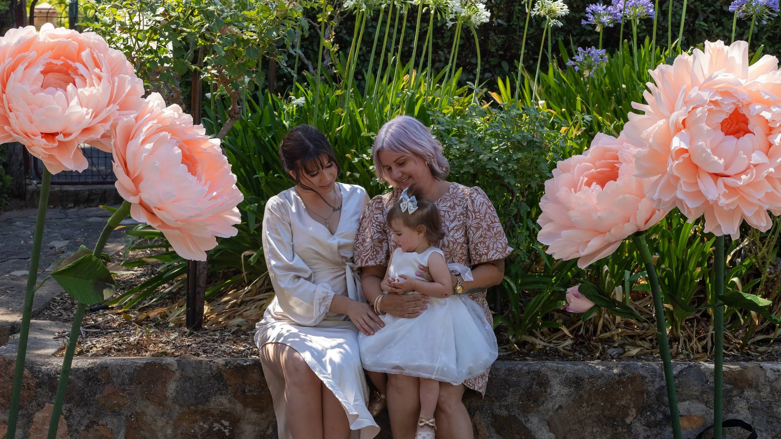 Three people, two women and a young girl, sitting on a stone wall in a garden with large pink peony flowers around them. The women are smiling and looking at the girl, who is holding a small object.