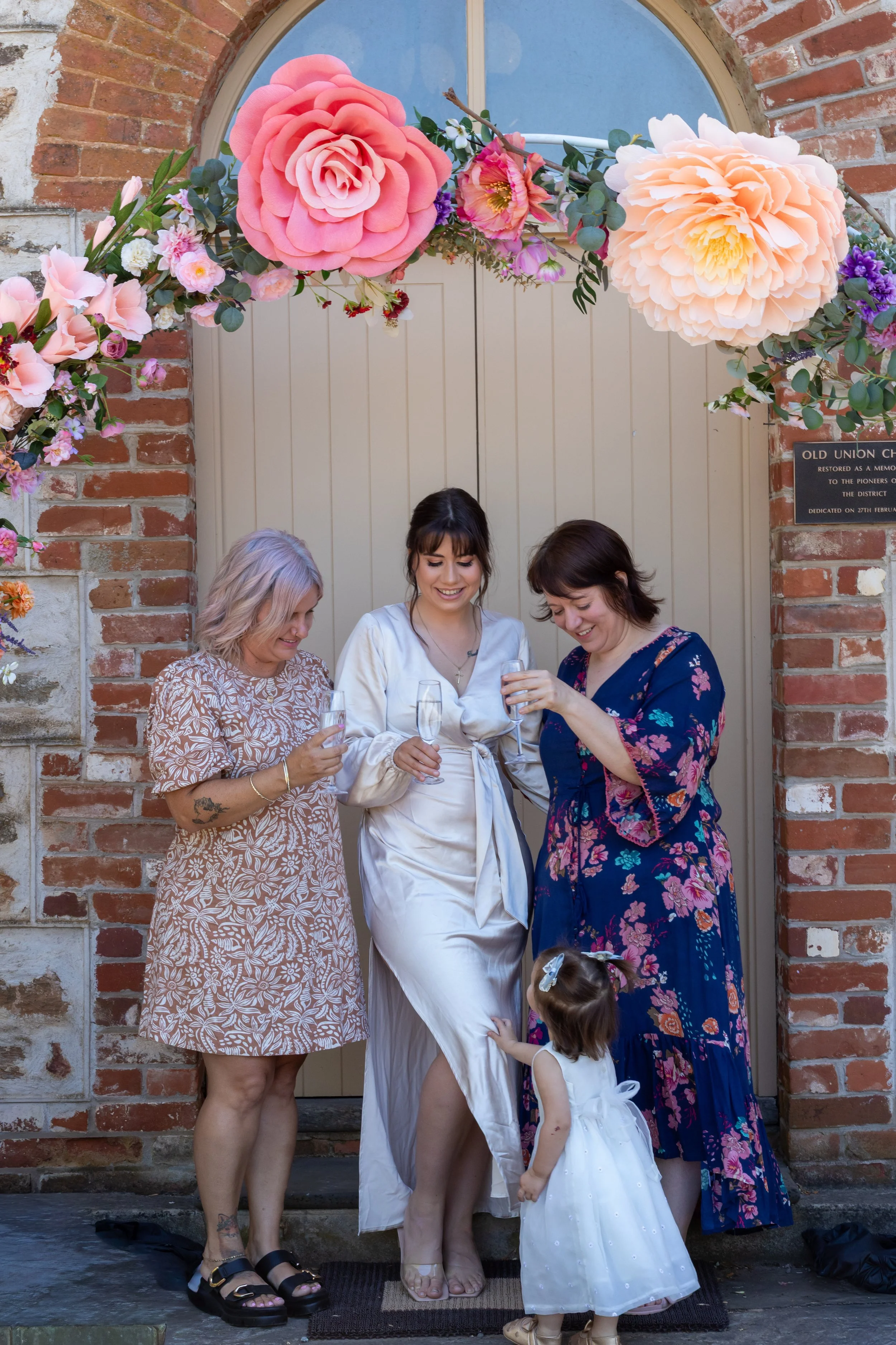 Three women and a young girl smiling and holding glasses of champagne standing in front of a decorated arch with large pink and peach paper flowers outside a brick building.