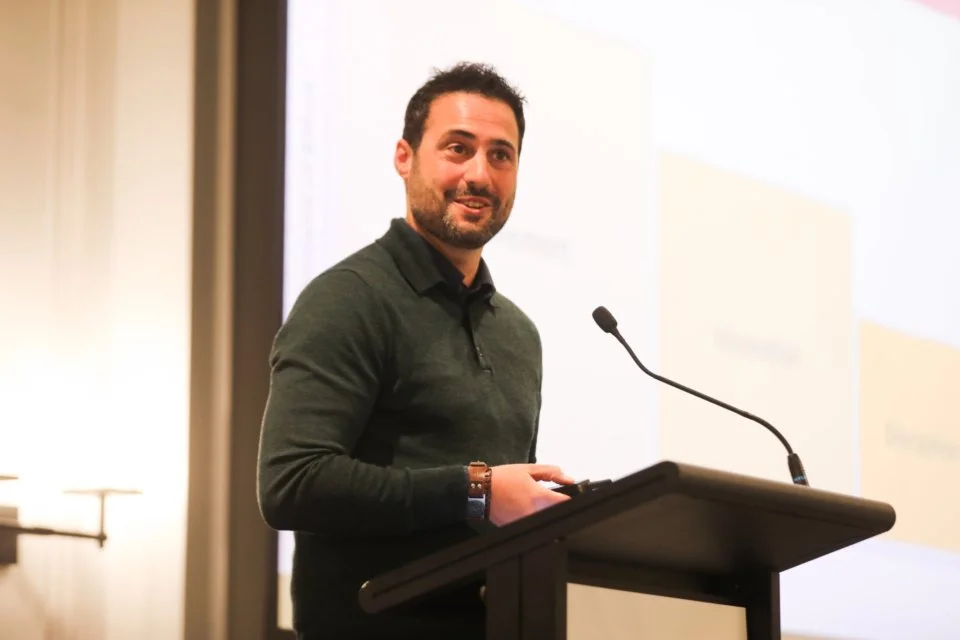 A man with dark hair and beard in a green sweater standing at a podium with a microphone, smiling, during a presentation.