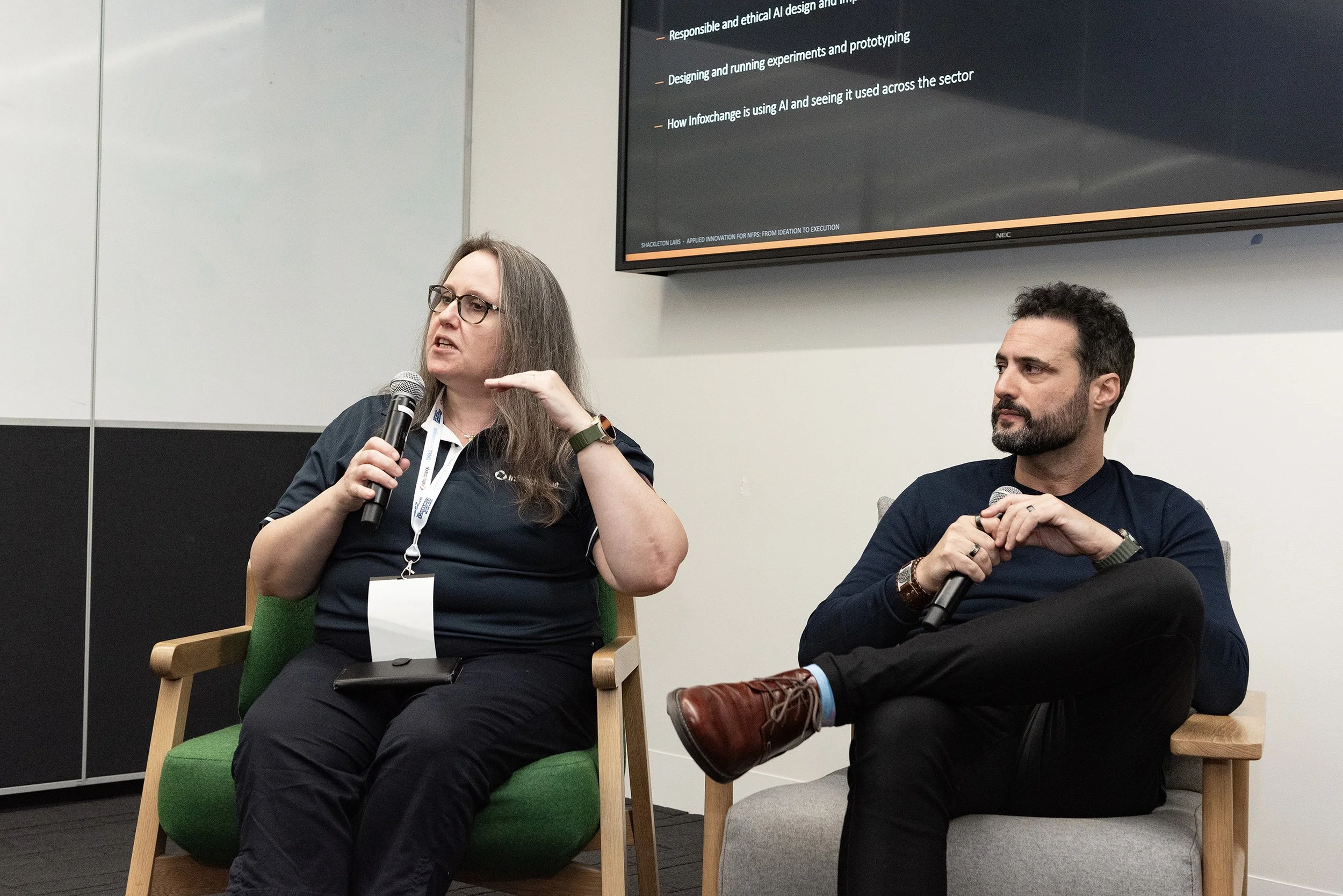 Two people seated in chairs participating in a panel discussion or presentation. The woman on the left is holding a microphone and speaking, wearing glasses, a dark polo shirt, and a lanyard. The man on the right is also holding a microphone, wearing a dark sweater, and has a beard. There is a large screen behind them displaying presentation slides with text related to artificial intelligence.