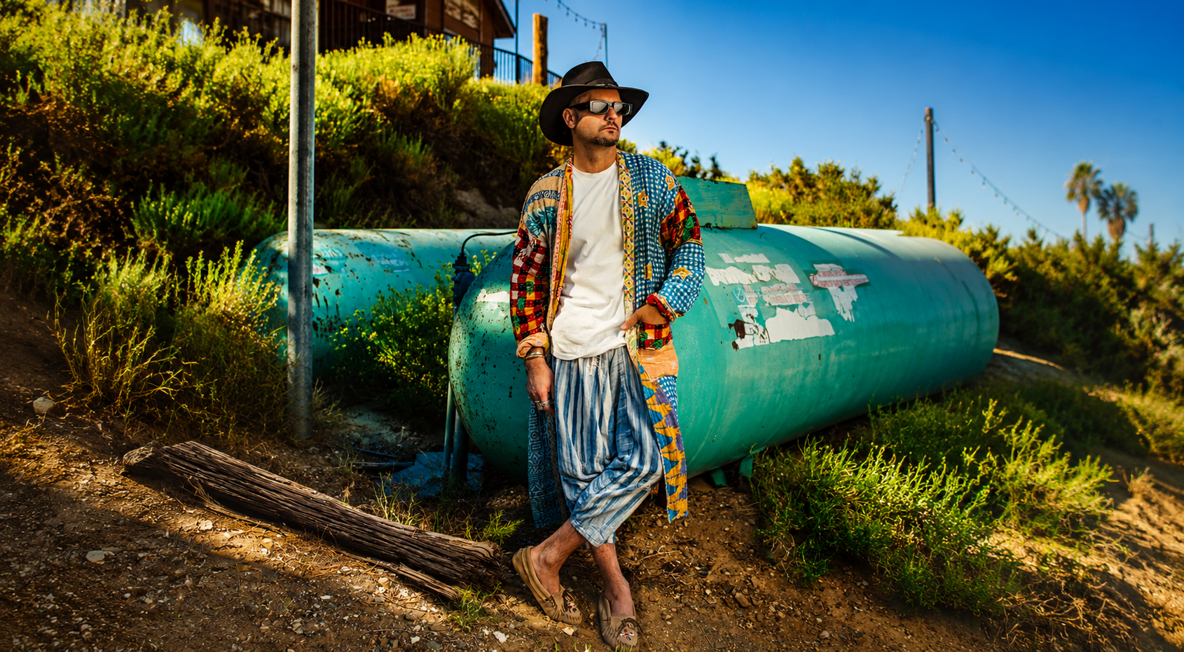 A man wearing a black wide-brimmed hat, dark sunglasses, a multicolored patchwork jacket, a white shirt, baggy striped pants, and slip-on shoes stands relaxed in front of a large, weathered green tank on a dirt path surrounded by green shrubs and bushes, under a clear blue sky.