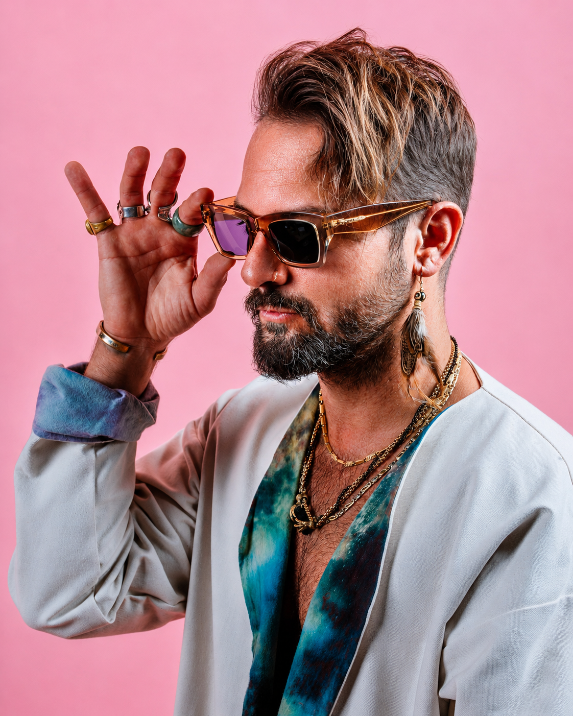 A man with styled hair and a beard is wearing sunglasses, multiple necklaces, earrings, and rings, posing against a pink background.