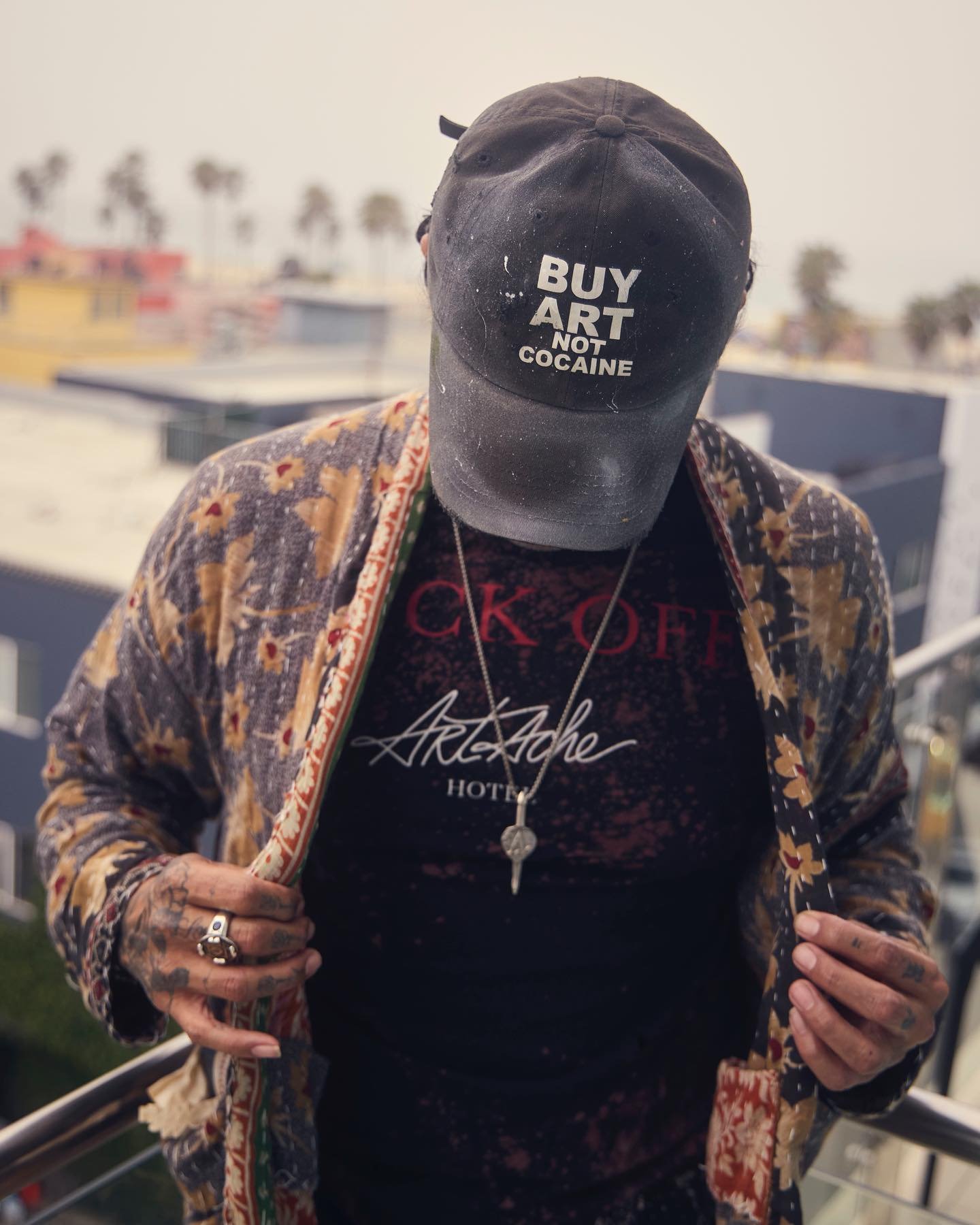 Person wearing a black cap with white text that says 'BUY ART NOT COCAINE' and a patterned jacket, standing outdoors with trees and buildings in the background.