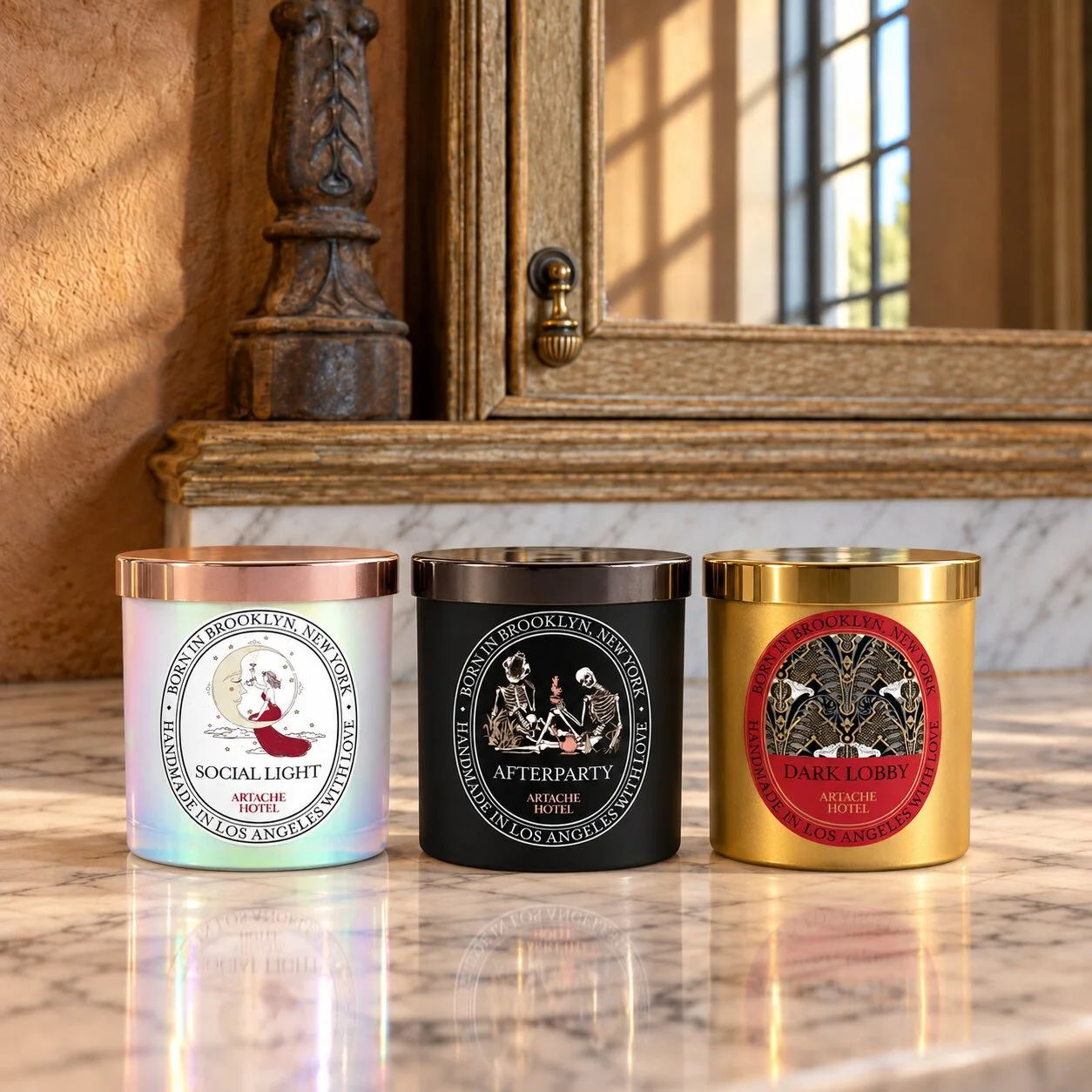 Three decorative candles in pink, black, and gold containers with vintage style labels and gold, copper, and black lids, placed on a marble surface with a wooden and mirror background.