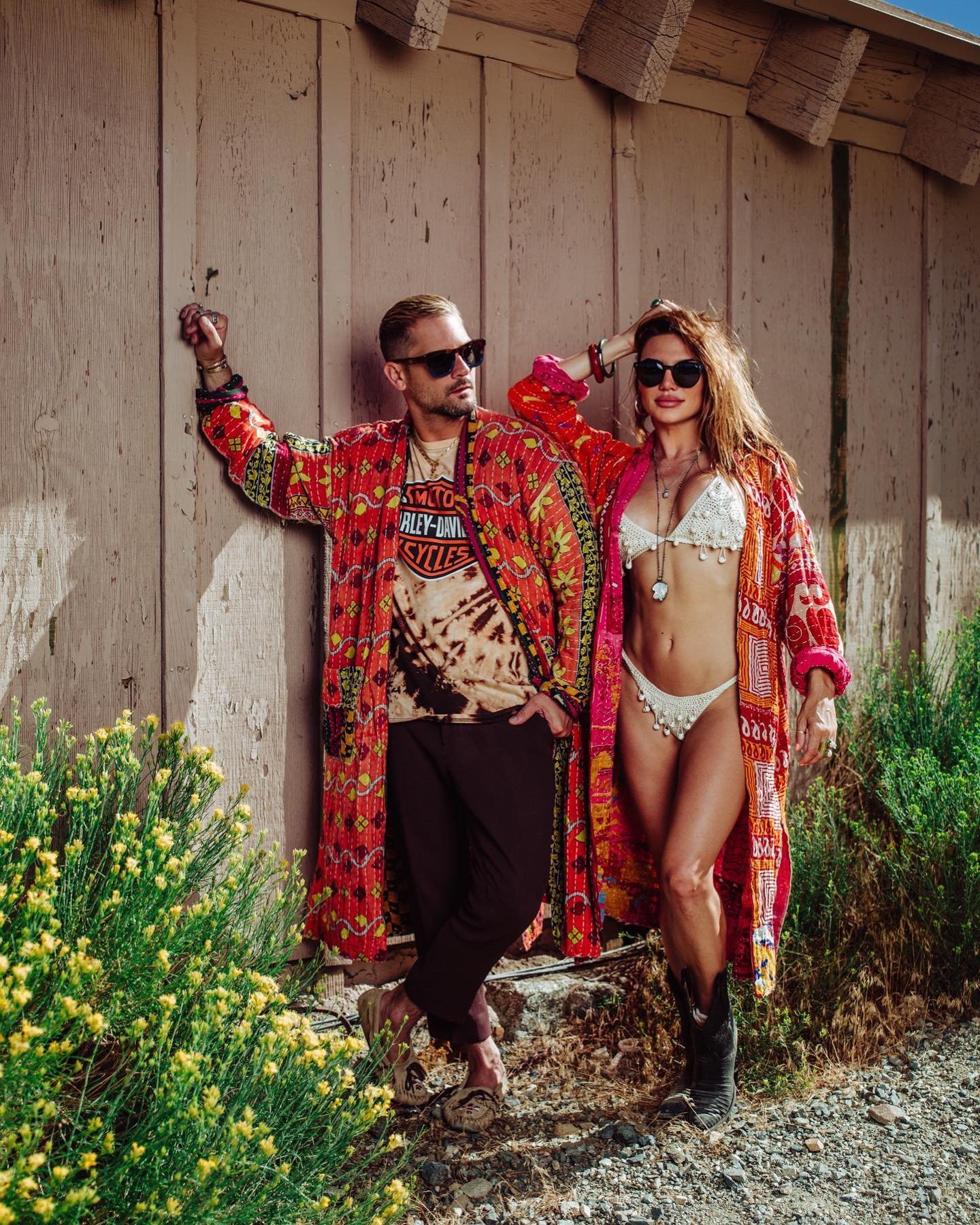 A man and woman posing outdoors against a wooden wall, both wearing sunglasses and vibrant, patterned clothing. The man has short hair, a beard, and is wearing a Harley Davidson shirt with a long, colorful coat and dark pants. The woman has wavy hair, wears a bikini with a kimono-style robe, and is accessorized with jewelry and black boots.