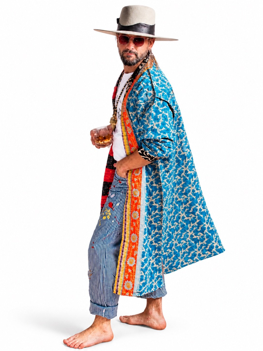 A man dressed in a colorful, eclectic outfit with a hat, sunglasses, and a drink in hand, standing barefoot and looking at the camera.