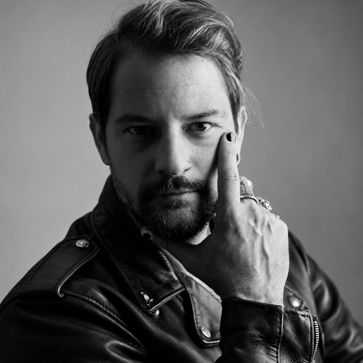 Black and white portrait of a man with a beard and mustache wearing a leather jacket, with his hand touching his face near his eye, looking directly at the camera