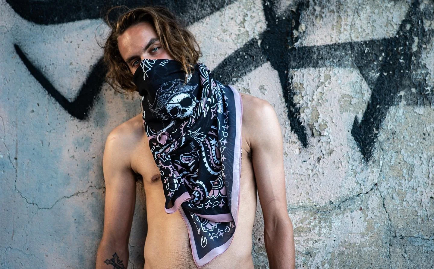 A shirtless young man with shoulder-length brown hair and light skin stands against a weathered, textured wall with graffiti. He wears a black bandana with white skull and star patterns, covering his nose and mouth. His right arm is tattooed with a star design.
