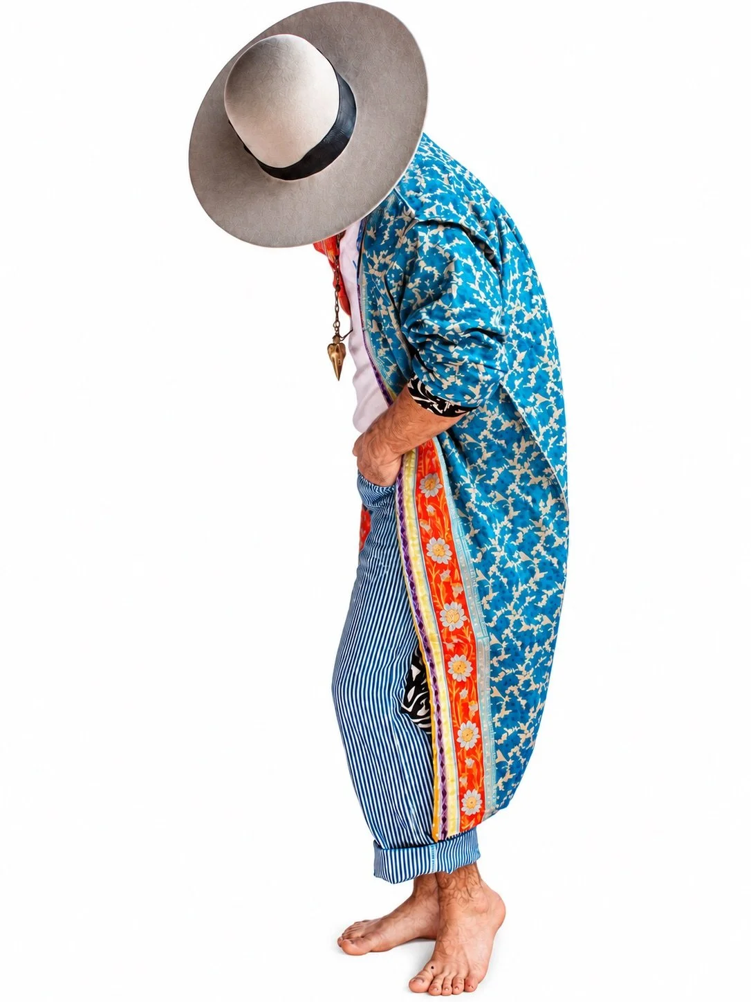 Person dressed in colorful, eclectic clothing with a large straw hat, standing barefoot against a white background.