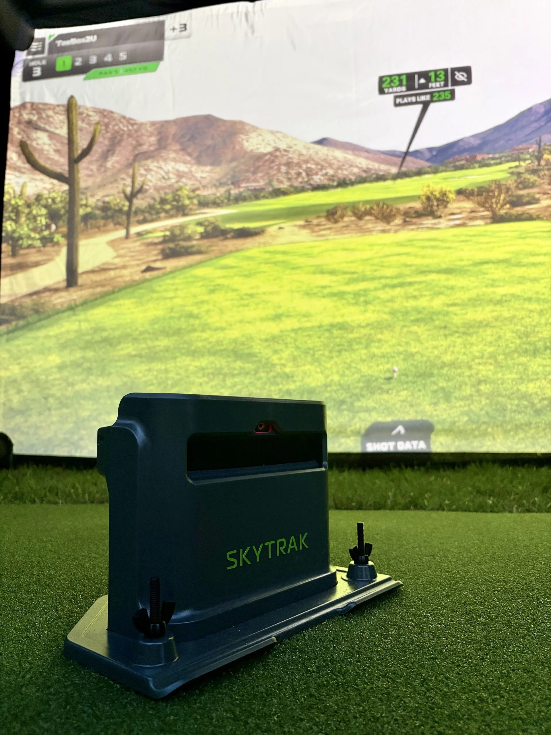 SkyTrak+ launch monitor on a hitting mat in front of the TeeBox2U golf simulator projection screen.