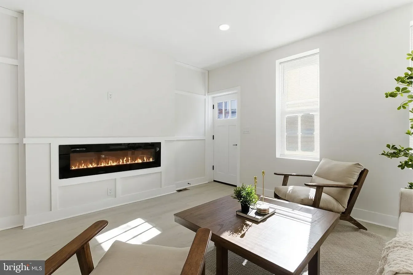 Bright living room with white walls, a modern electric fireplace, a window with blinds, a wooden armchair with beige cushions, a coffee table with a plant and decor, and a door leading outside.