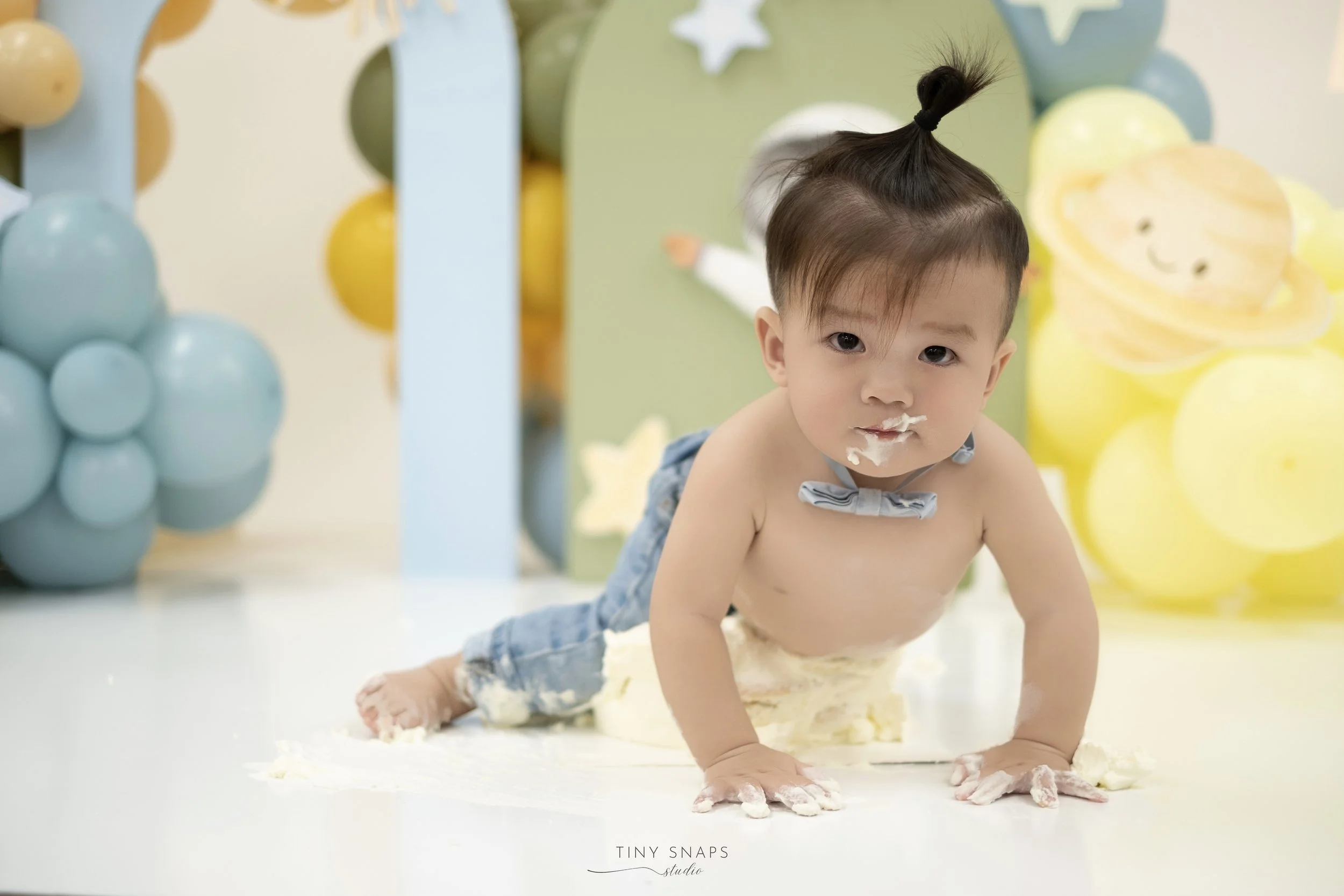 A baby crawling on the floor, with cake and frosting on their face and hands, surrounded by yellow and blue balloons and decorations.