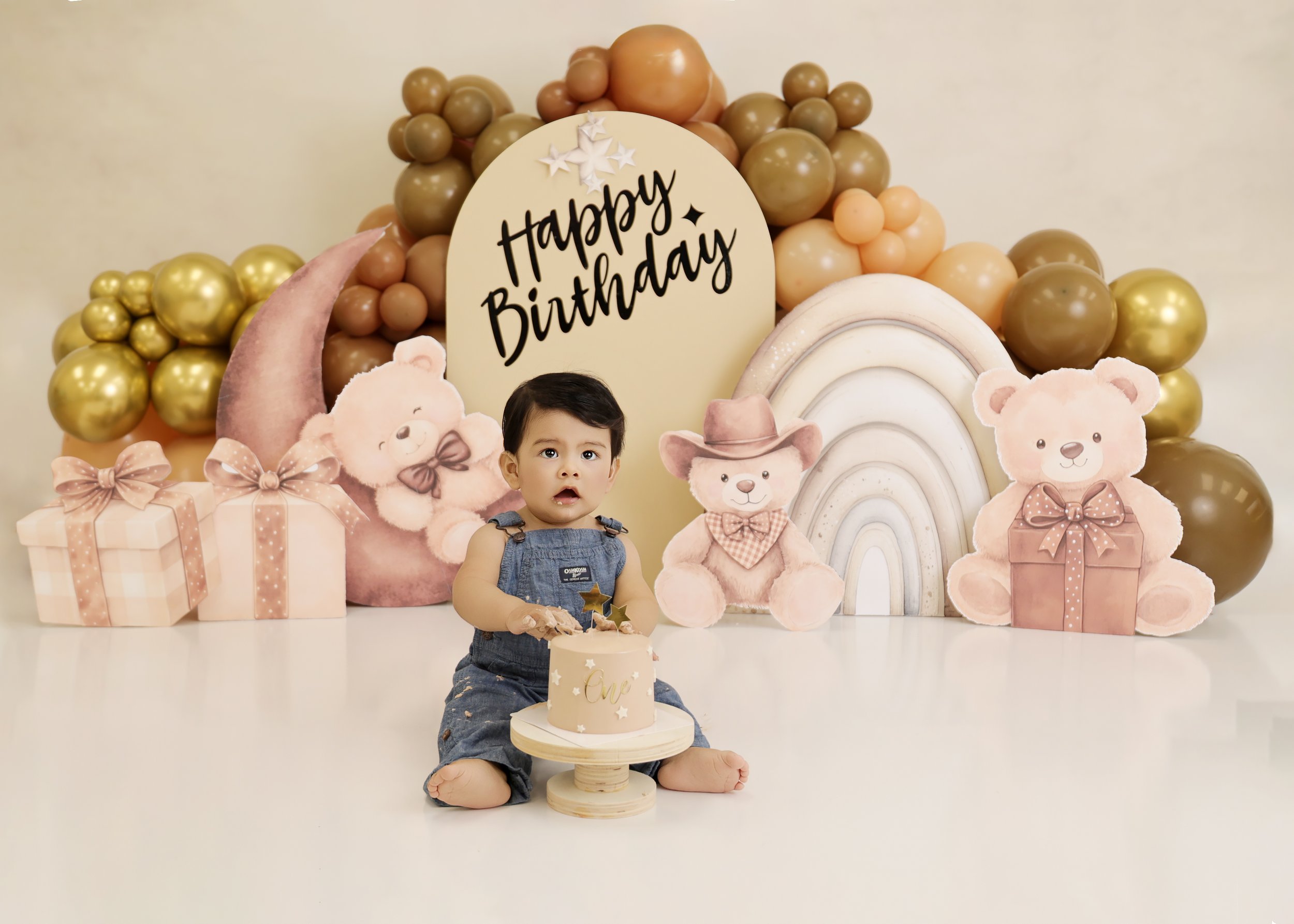 A young child sitting on a white floor holding a small cake with a gold star topper, surrounded by birthday decorations including teddy bear and rainbow cutouts, balloons, and a sign that says 'Happy Birthday' in black letters.