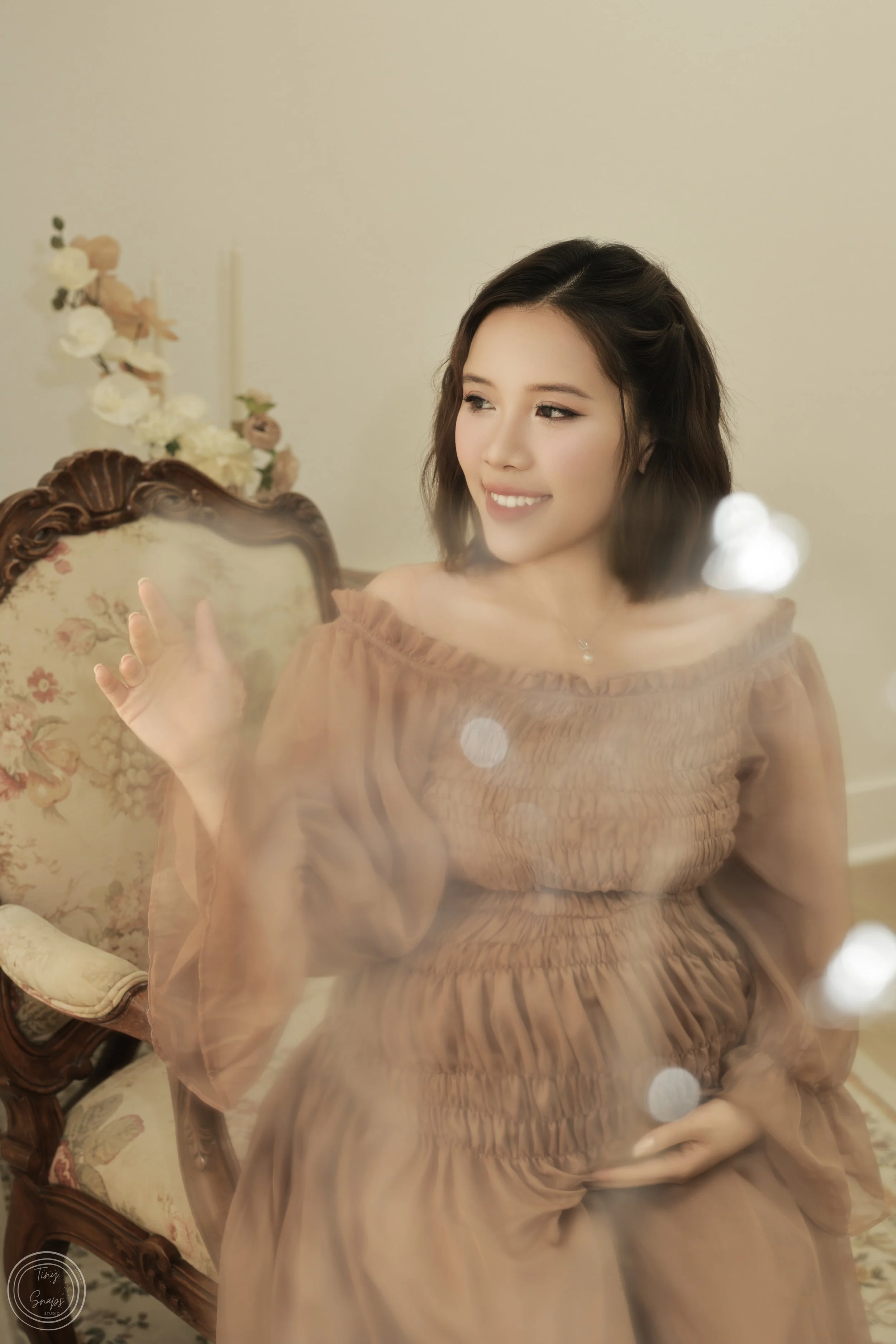 A young woman with shoulder-length dark hair smiling and looking to her right, sitting on an ornate floral-patterned vintage chair with a floral arrangement in the background, wearing a light brown off-shoulder dress.