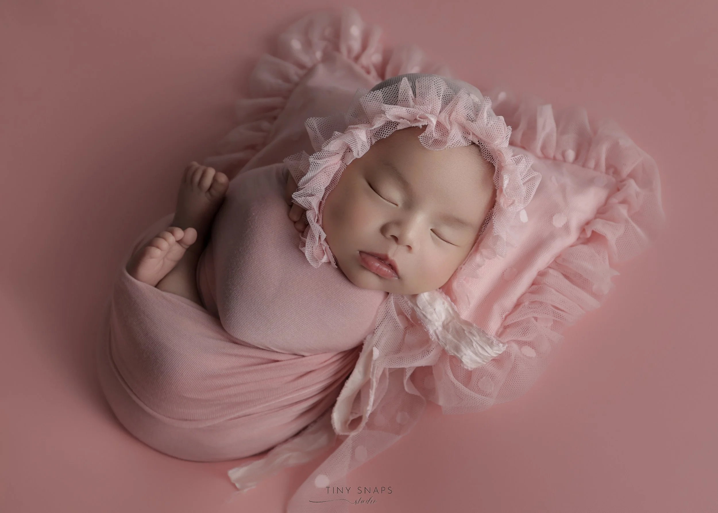 A sleeping baby girl wrapped in pink swaddle blanket, wearing a pink bonnet with ruffles, lying on a pink surface with ruffled pillow.