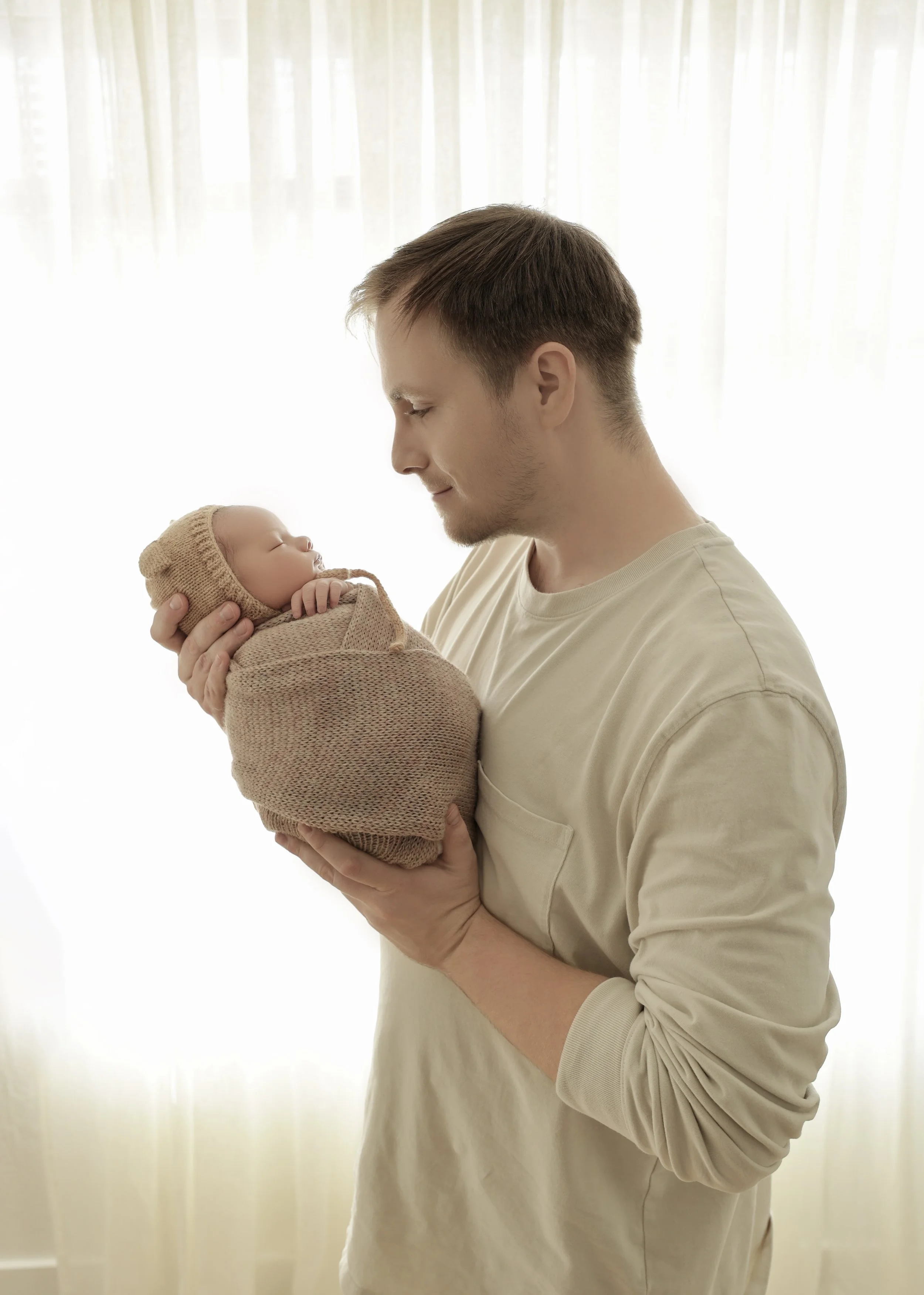 A young man holding a sleeping baby wrapped in a beige knit blanket, with a matching hat, against a light background with sheer curtains.