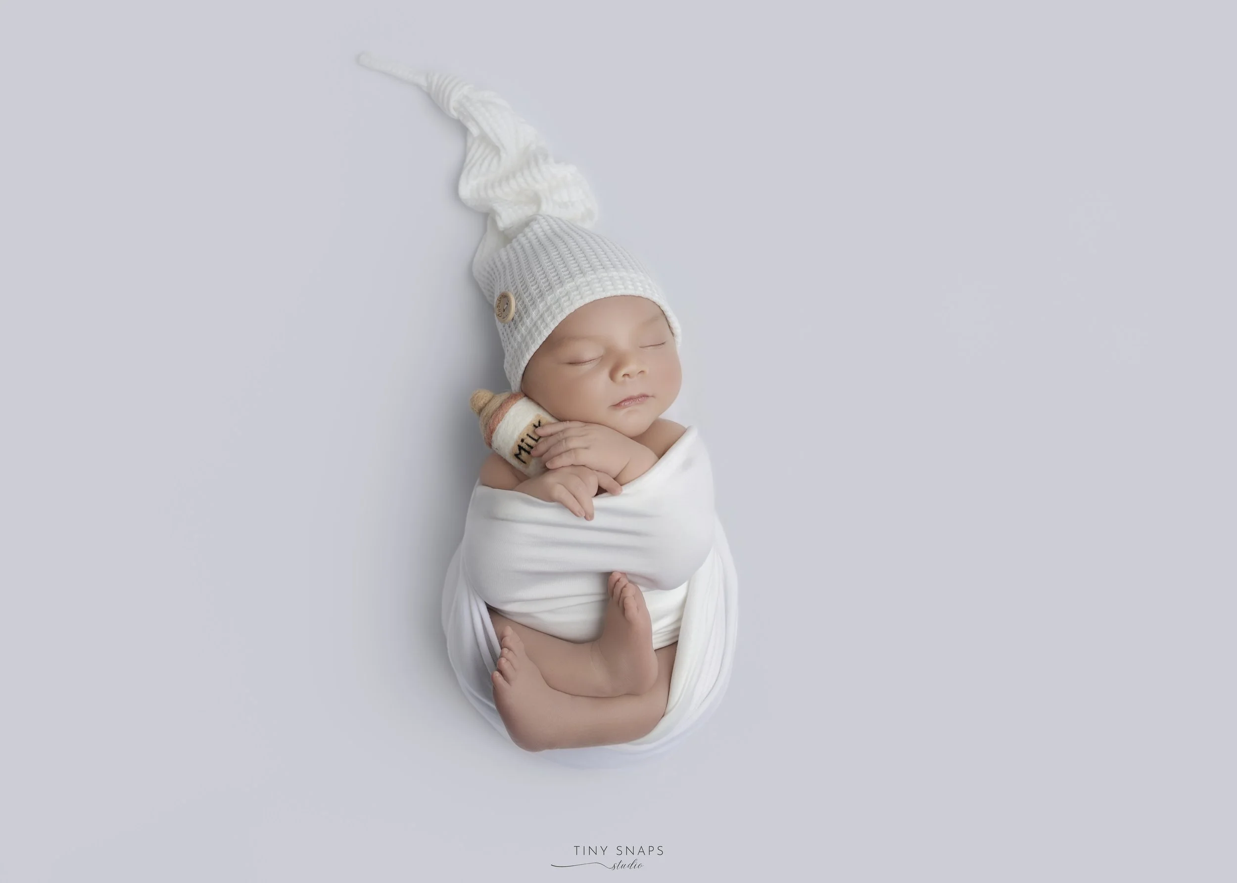 Adorable sleeping baby wrapped in white cloth, wearing a white knitted hat with a long, pointed tail, holding a small plush milk bottle, set against a light gray background.