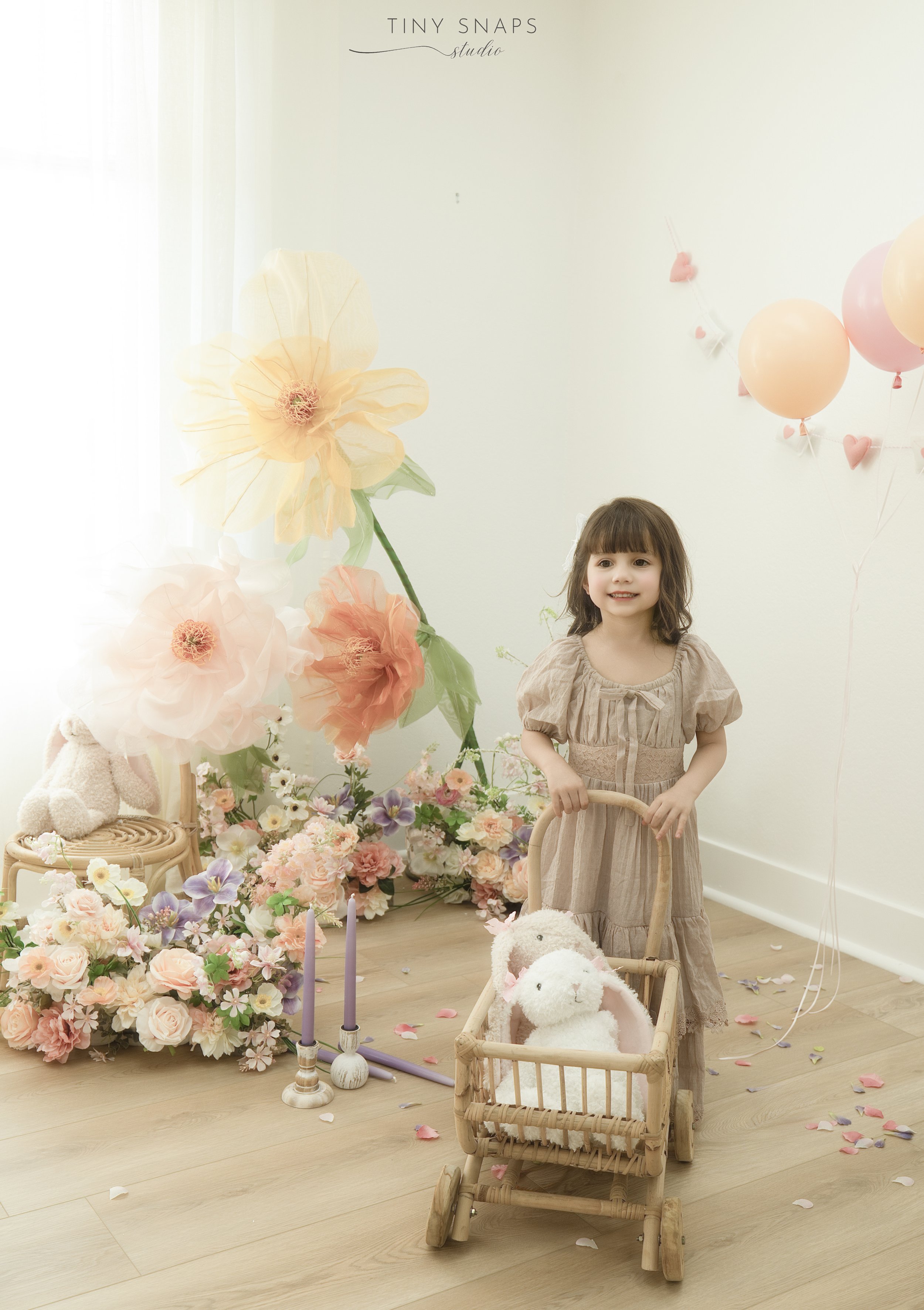 A young girl standing indoors, holding a toy stroller with plush bunny toys inside, surrounded by pastel-colored flowers, balloons, and decorative items, in a softly lit room with a white wall.