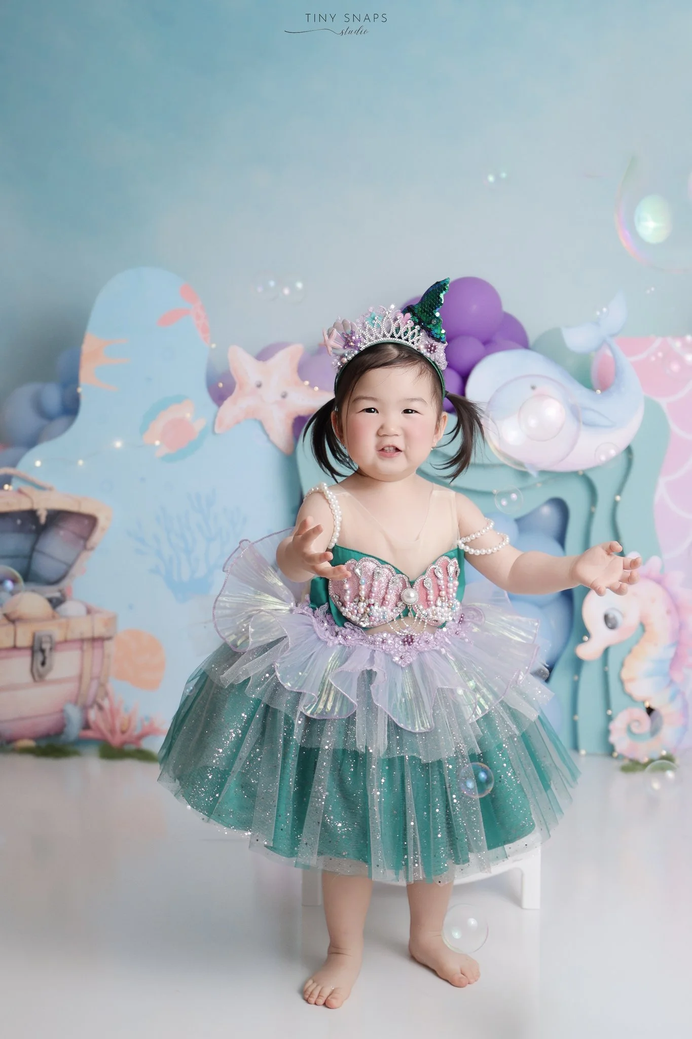 Child in a colorful mermaid costume with a crown, standing in front of an ocean-themed backdrop with sea creatures and treasure chest.