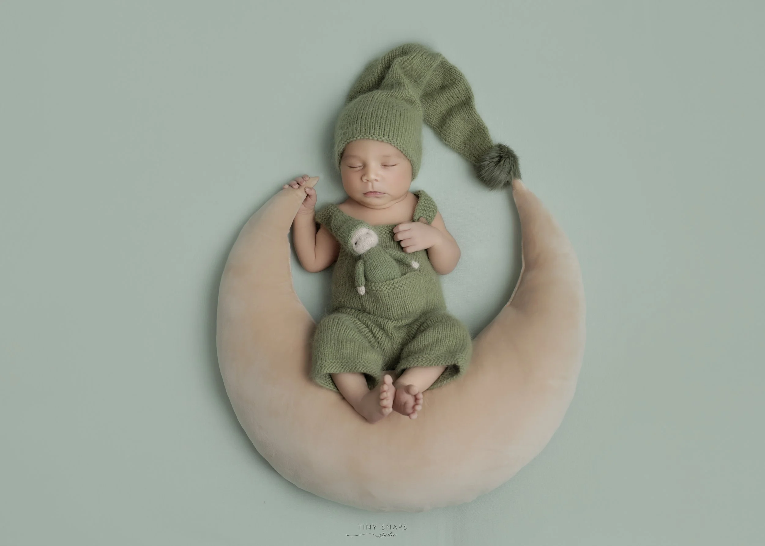 A newborn baby dressed in green knit clothing with a hat and suspenders, sleeping on a crescent-shaped moon pillow, holding an adult's finger. The background is a pale green wall.