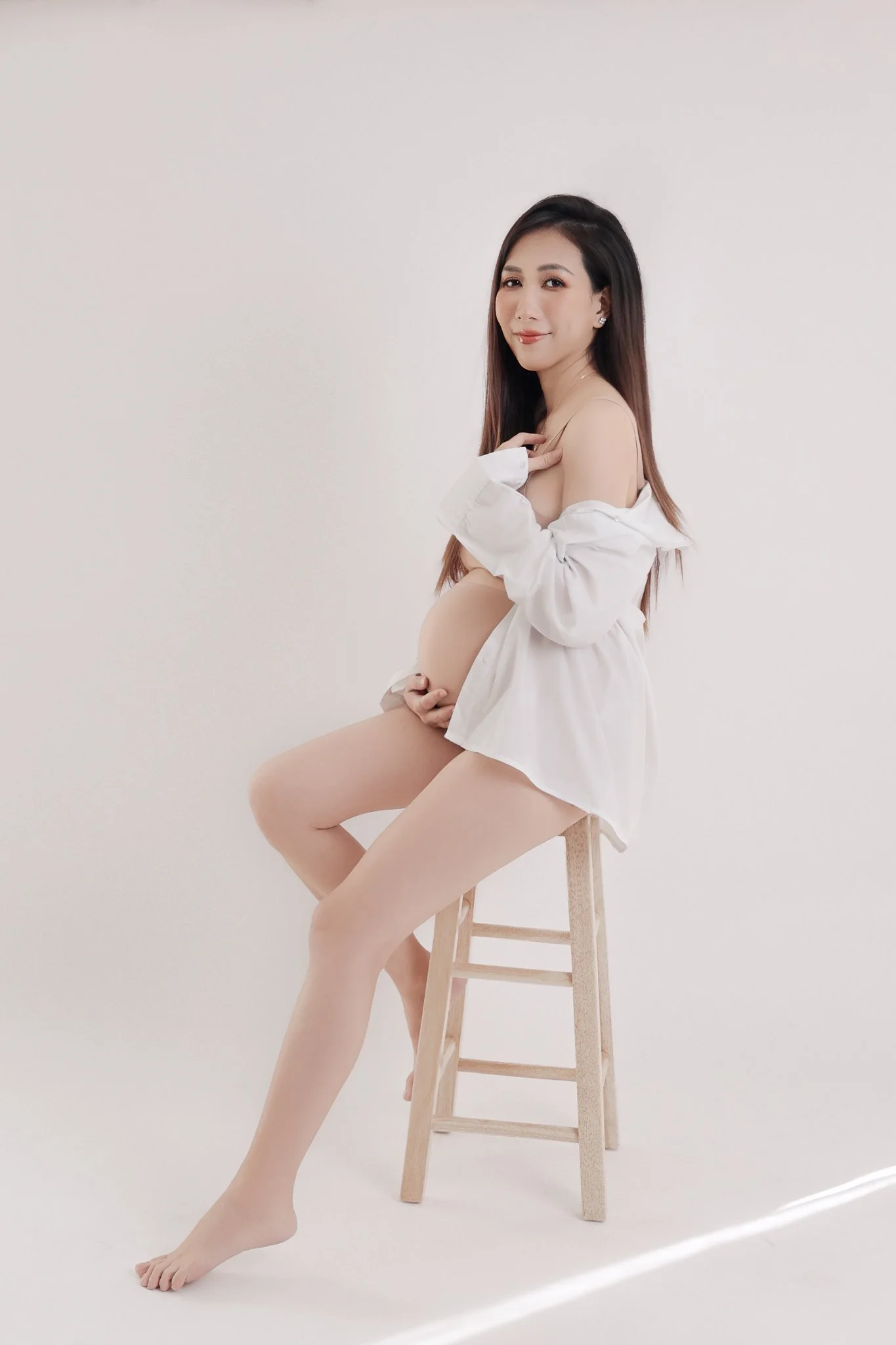 A woman with long dark hair, sitting on a wooden stool, dressed in a white shirt and nude stockings, against a plain light background.
