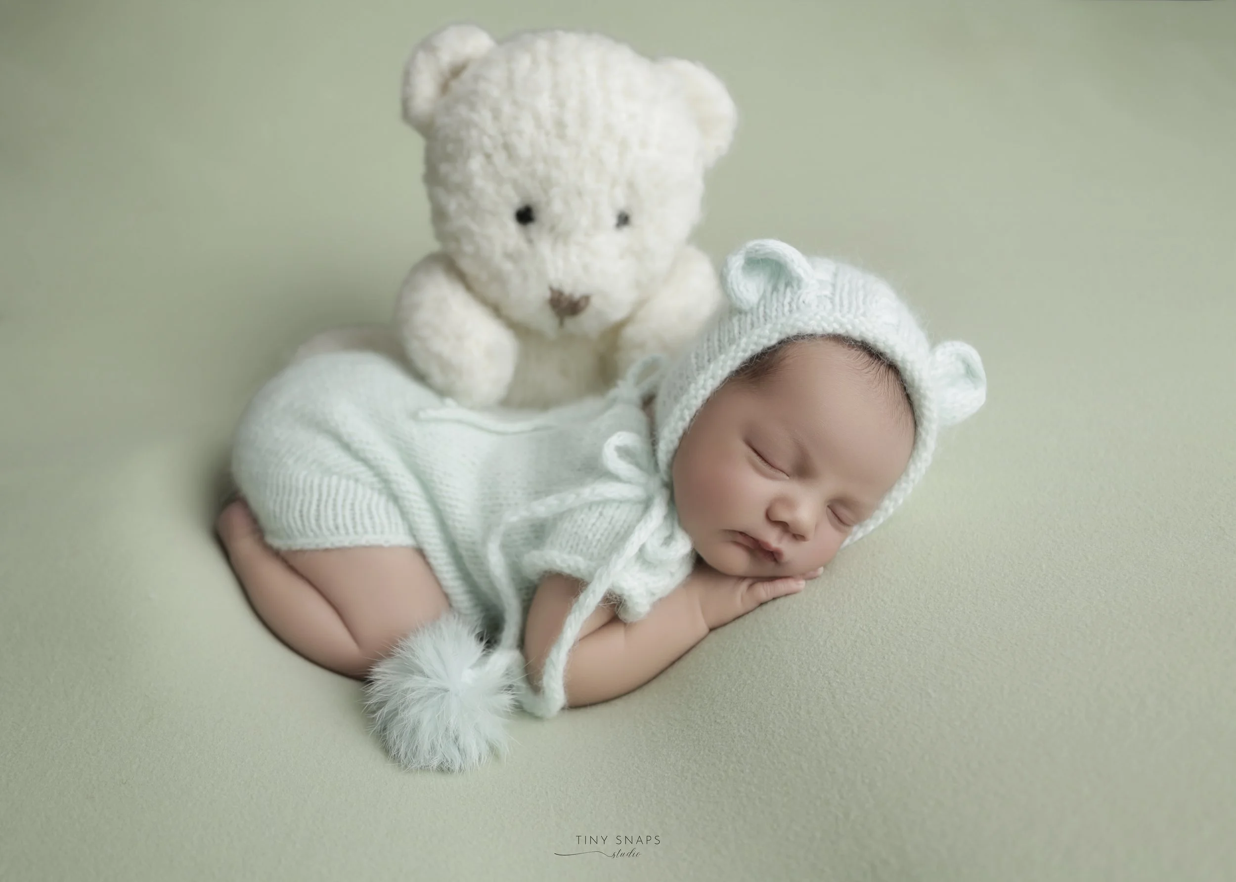 Sleeping baby wearing a knitted hat with bear ears, lying on a soft surface beside a plush white teddy bear.