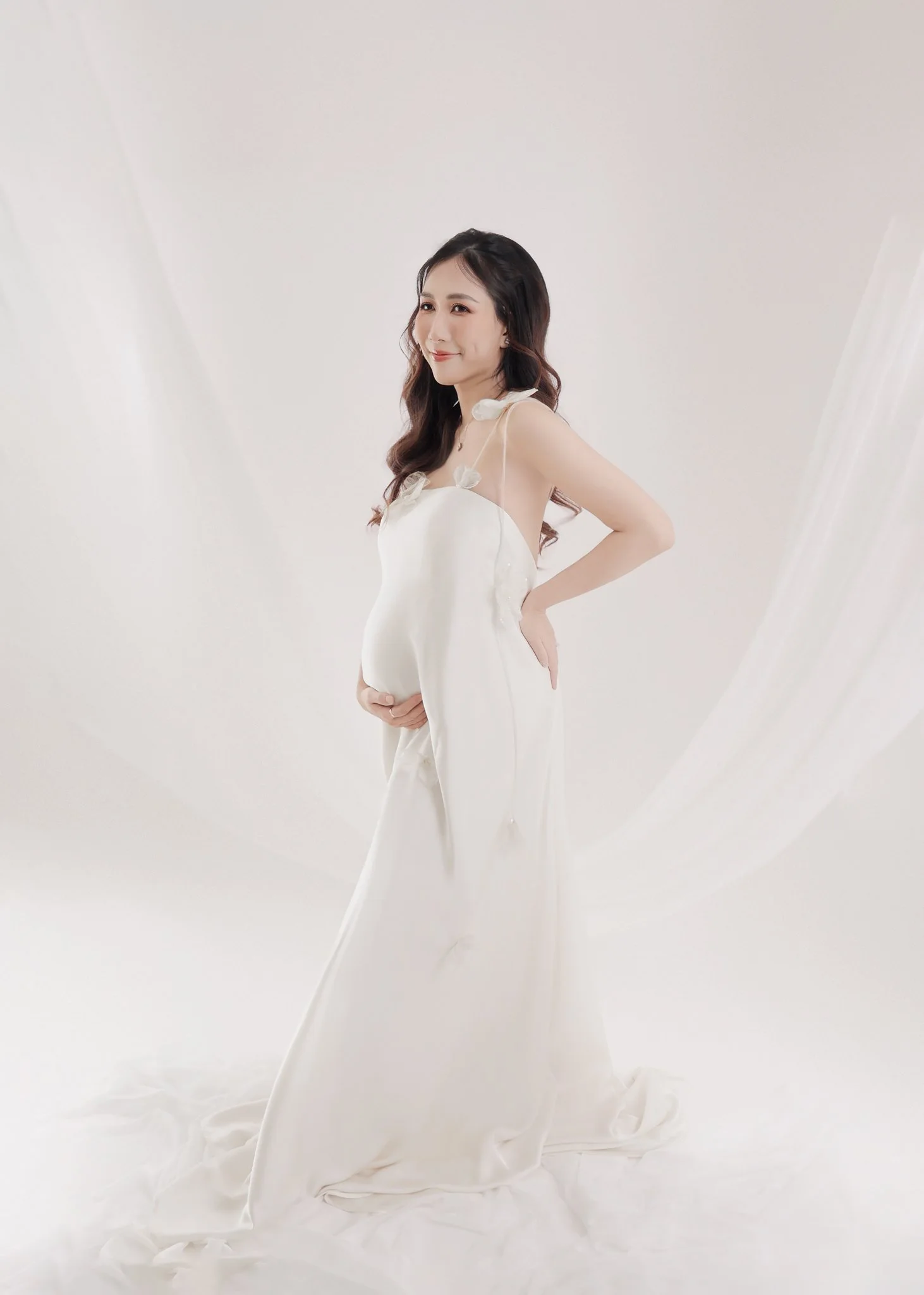 A pregnant woman in a white dress standing in a bright, minimal studio with white drapes.