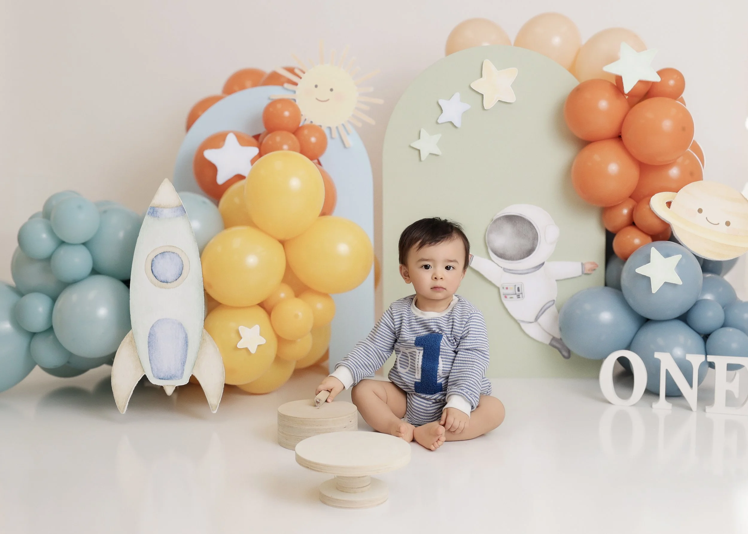 A young child sitting on the floor during a celebration with space-themed decorations, including balloons, a rocket cutout, space aliens, a smiling sun, and a large 'ONE' sign.