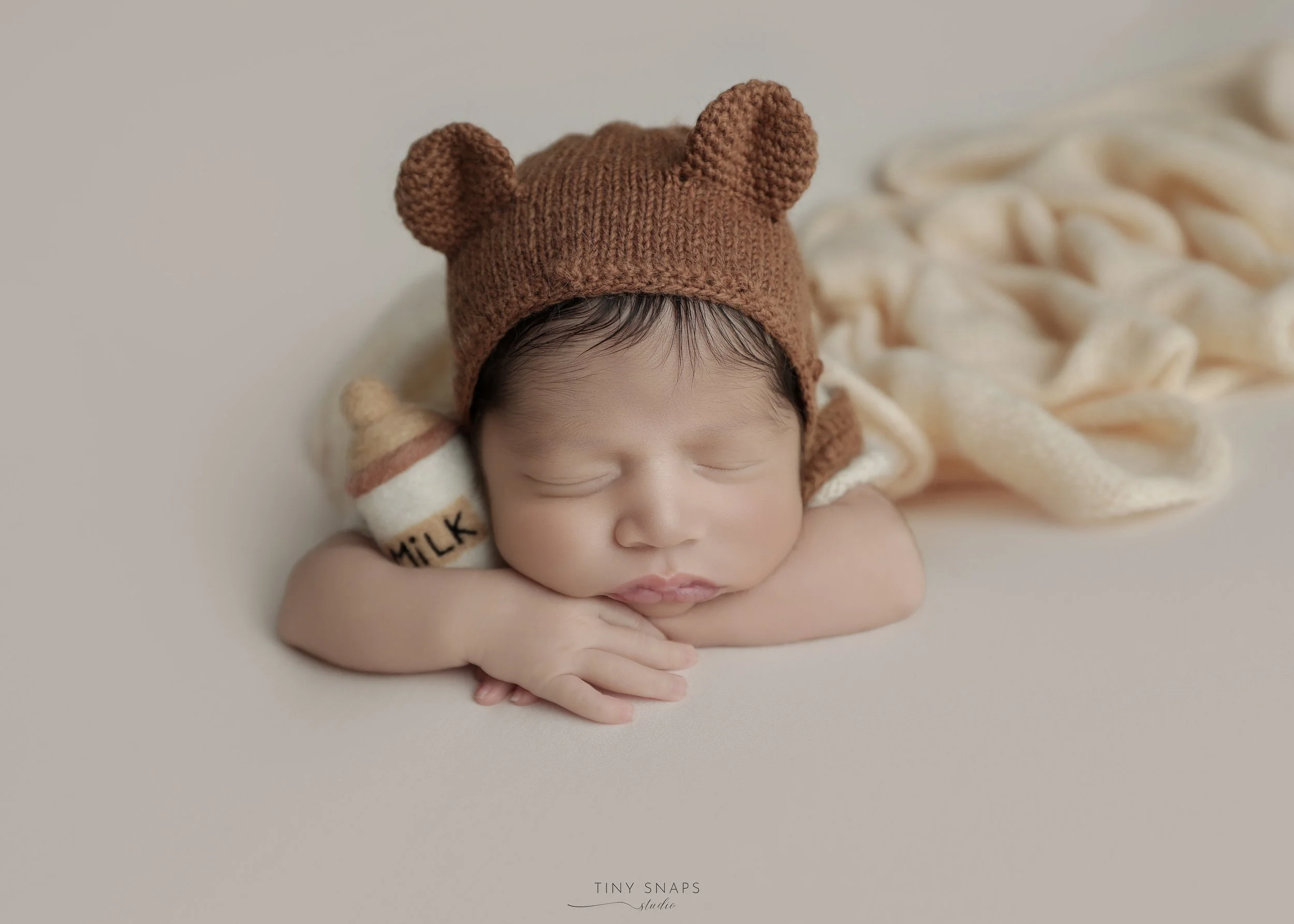 A sleeping baby wearing a brown knitted hat with bear ears, resting its head on its arms on a soft, cream-colored surface, with a small sock labeled 'milk' next to its face.