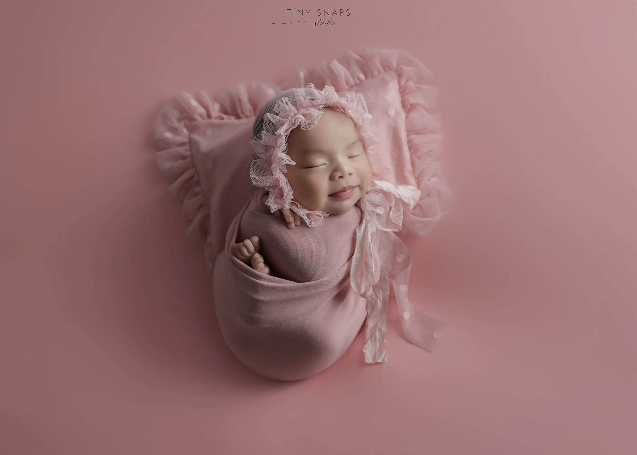 Smiling baby girl with a bonnet, wrapped in pink cloth, lying on a pink ruffled blanket, on a pink background.