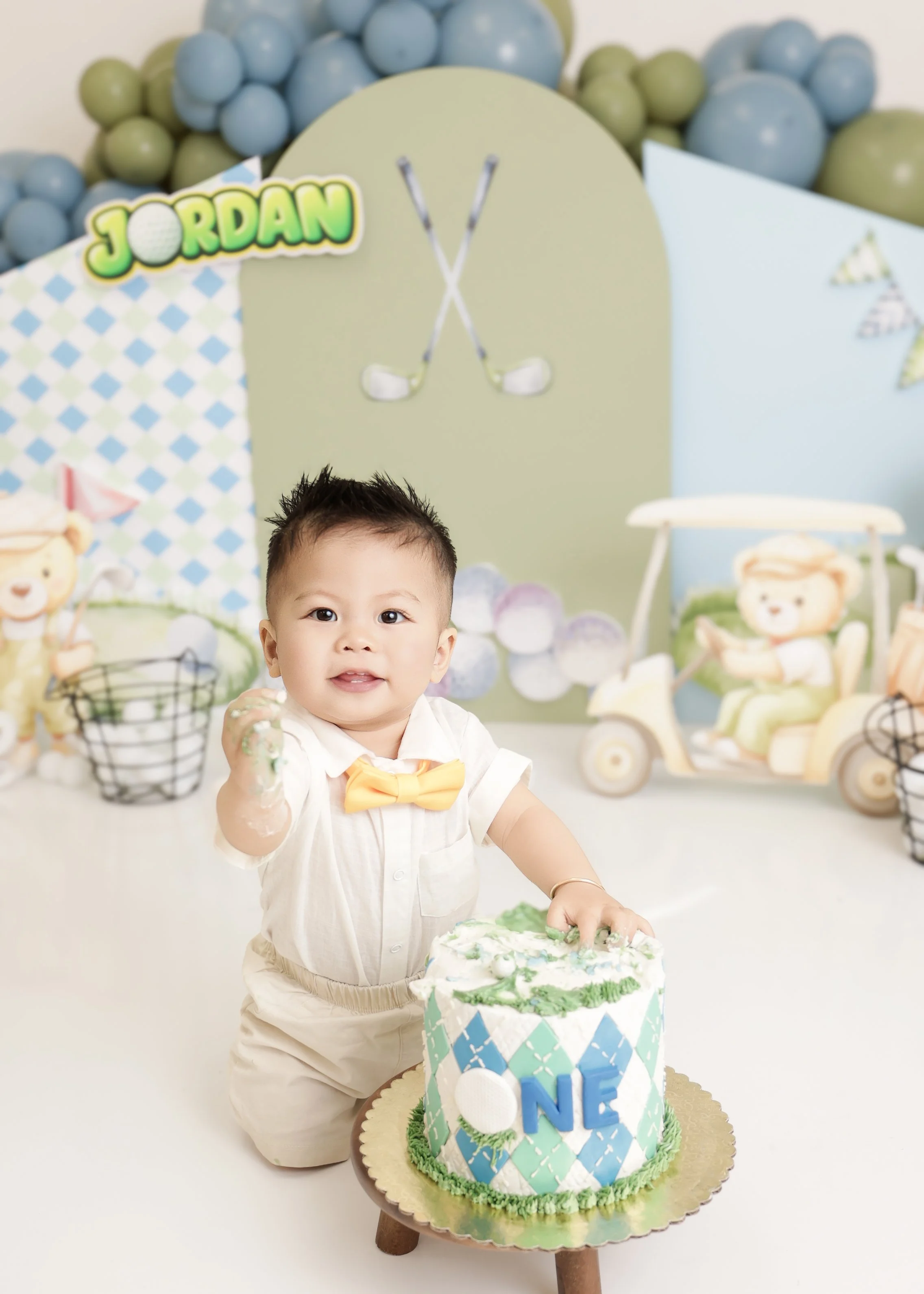 A young child at a birthday celebration, sitting on the floor with a cake in front, surrounded by decorations including balloons, a backdrop with golf motifs, and plush toys. The cake has the word 'ONE' on it, indicating the child's first birthday.