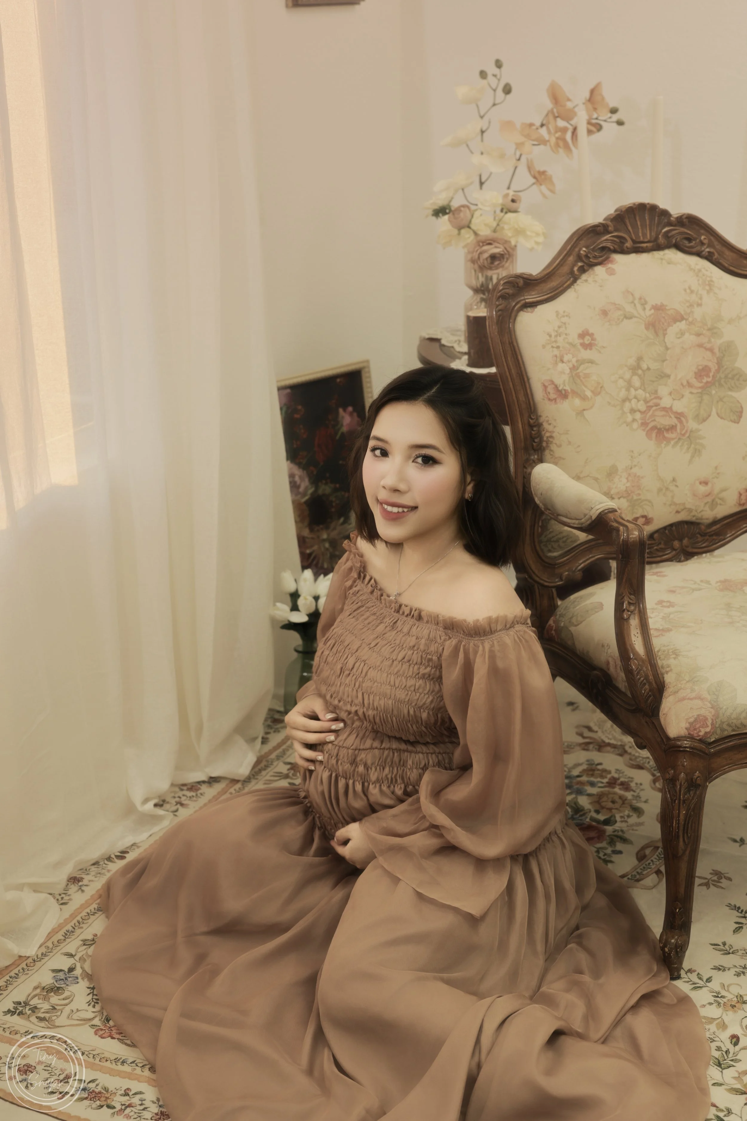 A pregnant woman sitting on the floor next to a vintage armchair, smiling at the camera, in a warmly decorated room with floral and vases.