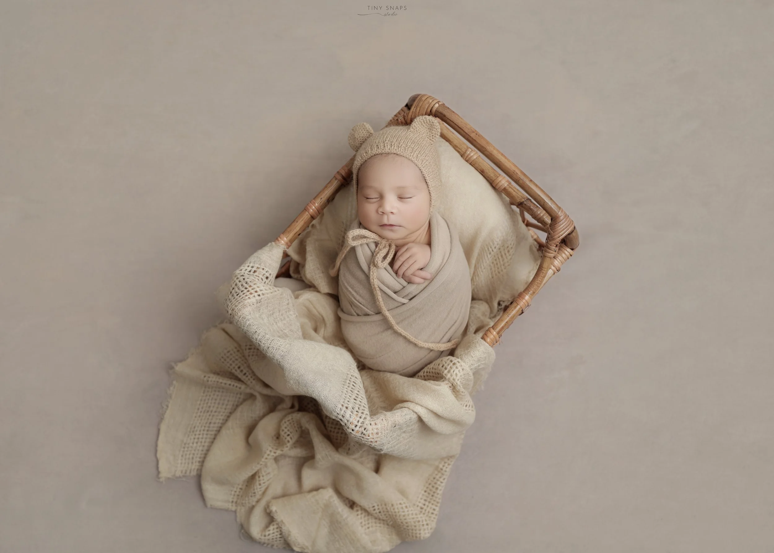 A sleeping baby in a woven basket, wrapped in beige cloth, wearing a knit hat with bear ears, surrounded by soft cream-colored blankets.