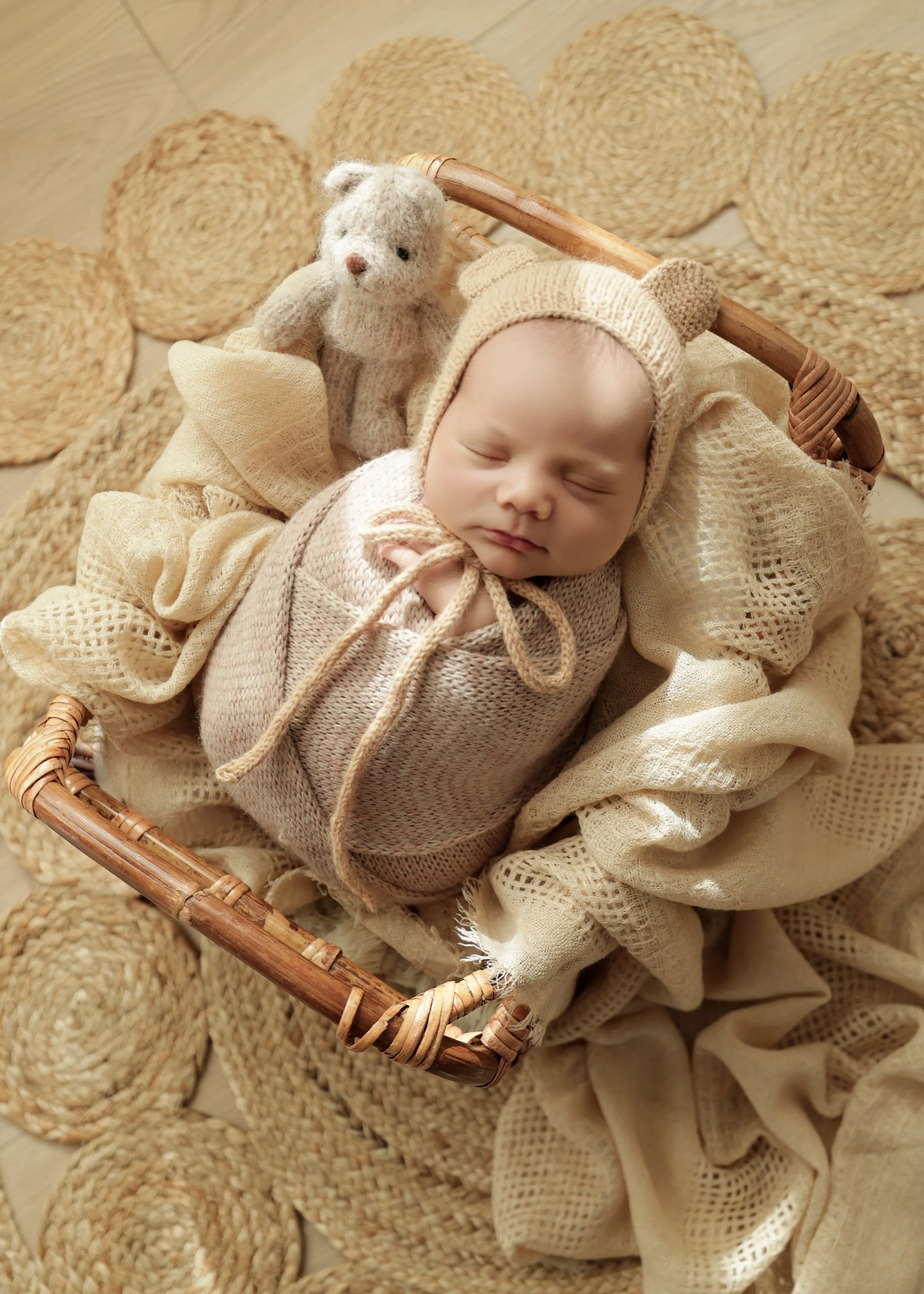 A sleeping baby wrapped in beige knitted clothing, wearing a bear hat, lying in a wicker basket, accompanied by a stuffed teddy bear, surrounded by beige woven mats and blankets.