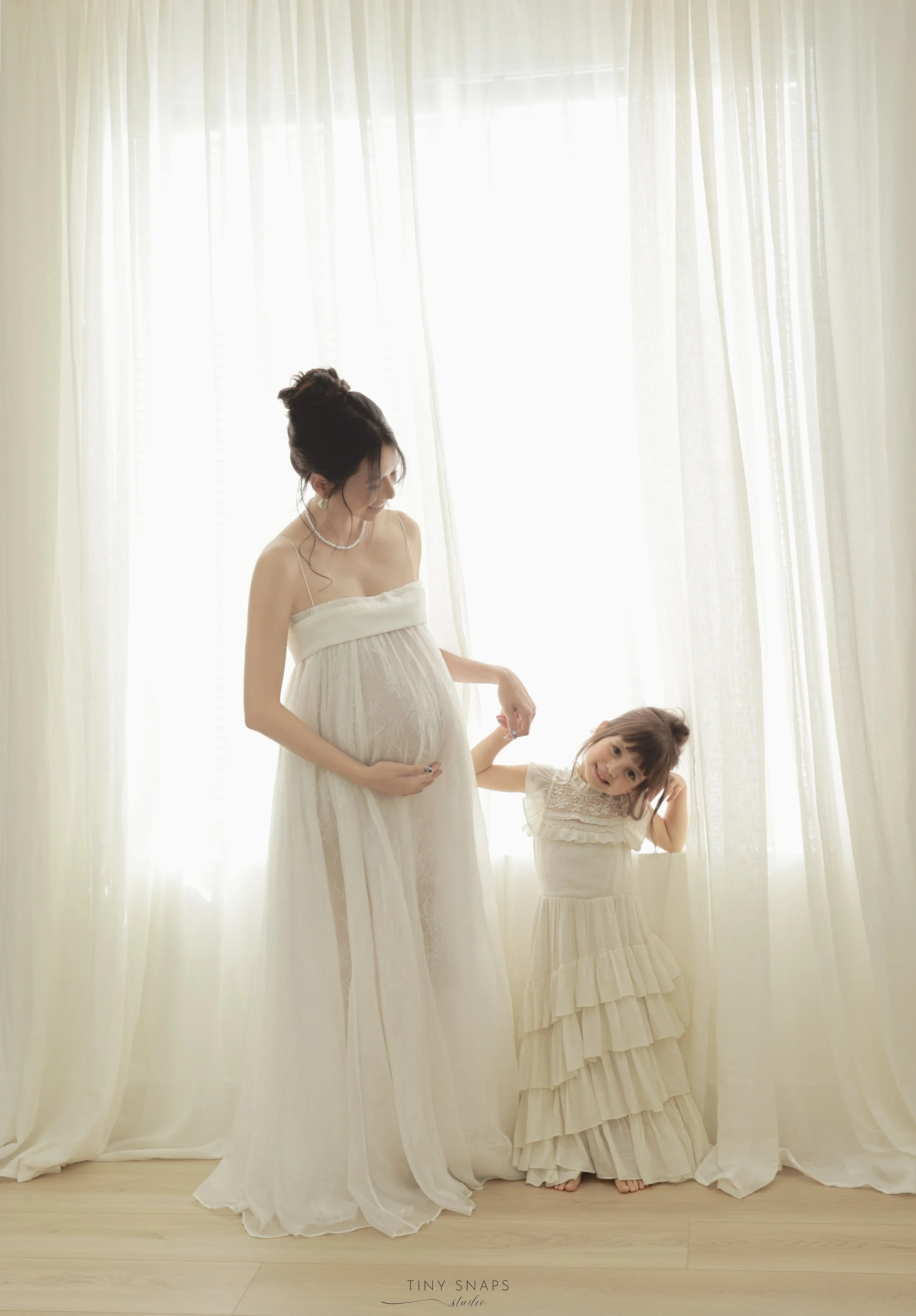 A pregnant woman and a young girl, both dressed in light-colored dresses, standing by a window with sheer curtains, holding hands and smiling.