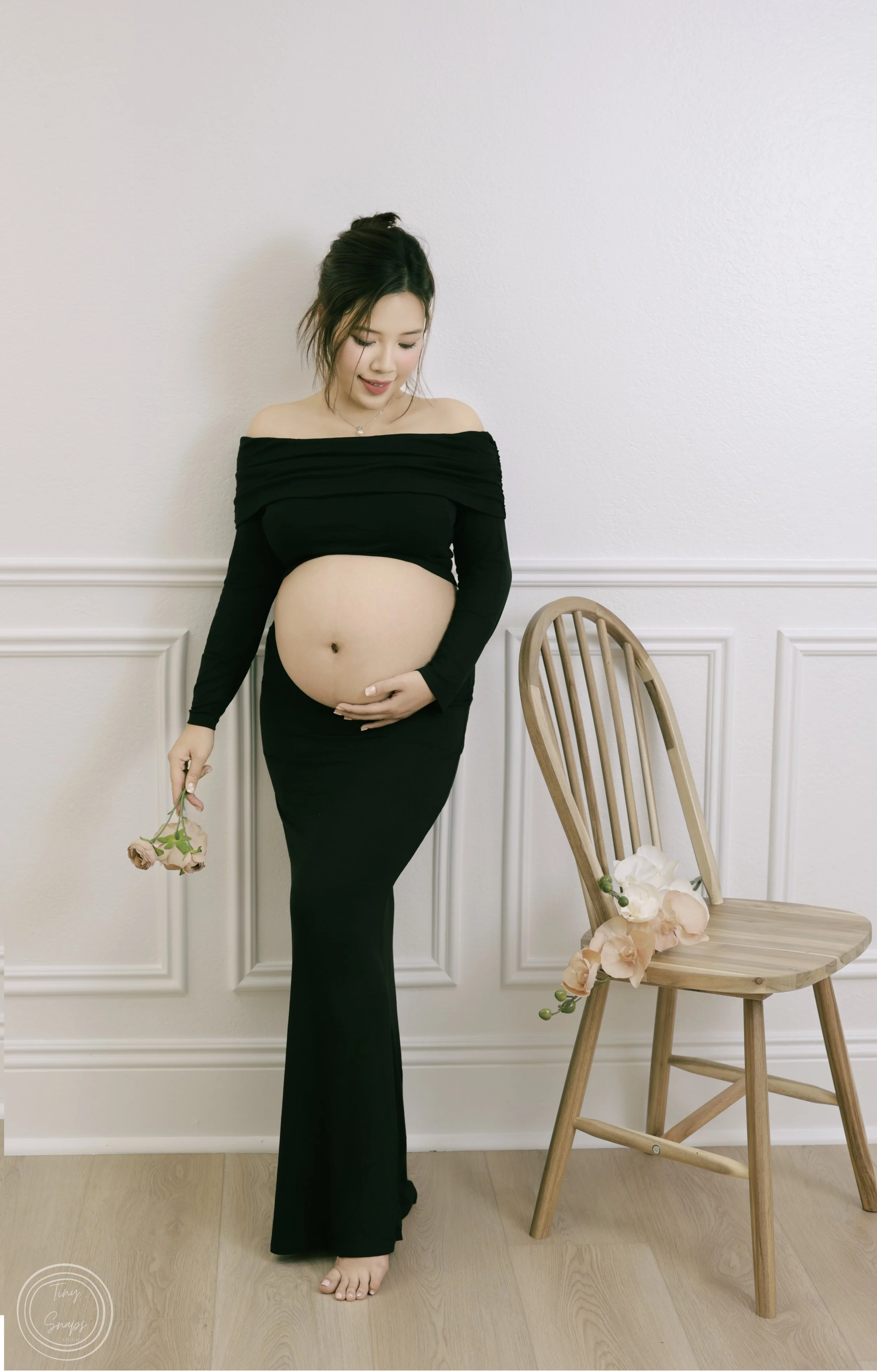 Pregnant woman in black dress standing barefoot indoors, holding flowers in one hand and touching her belly with the other, near a wooden chair decorated with flowers.