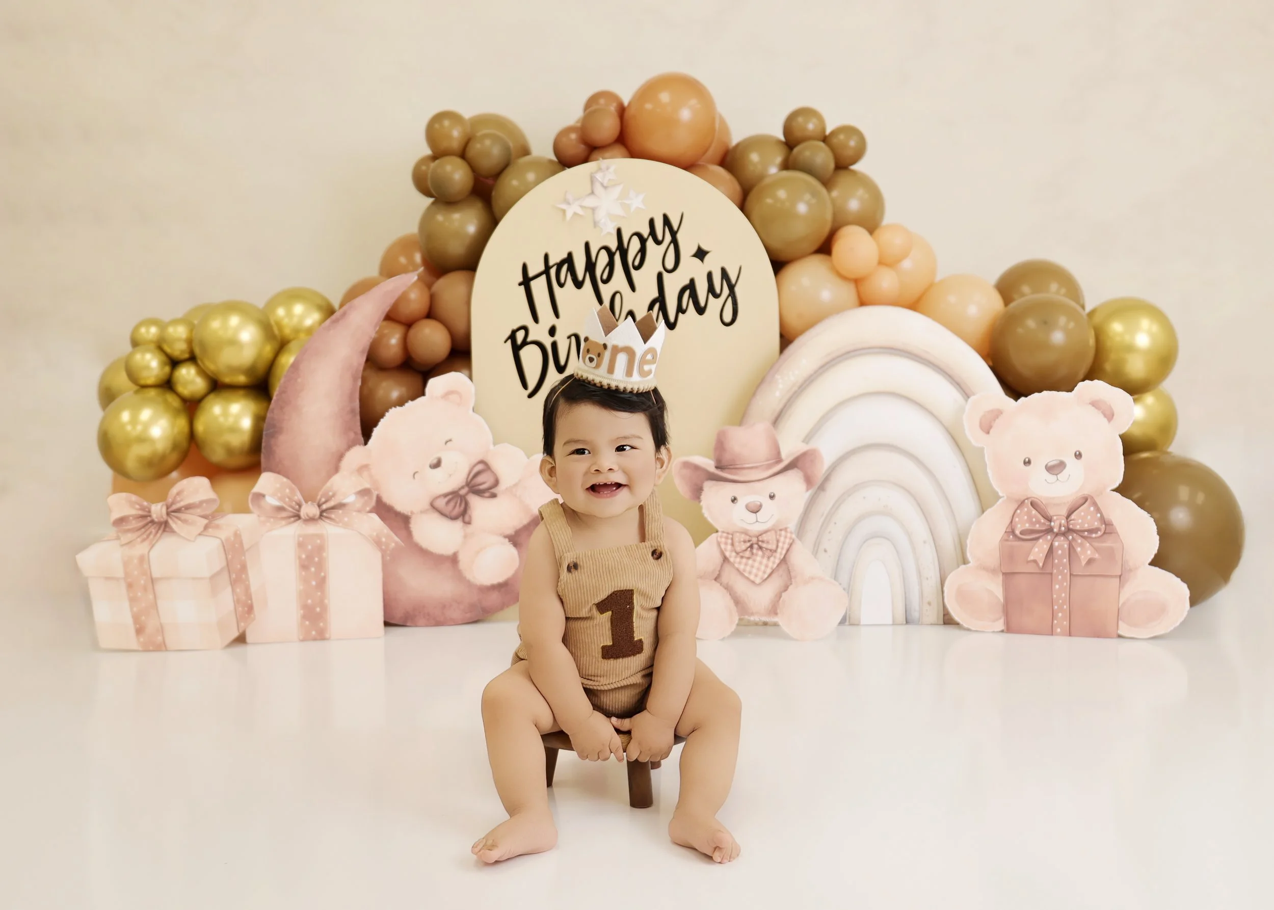 A smiling baby boy is sitting on a small stool in front of a birthday backdrop. The backdrop features balloons, plush teddy bears, a rainbow, and a moon with a banner that reads "Happy Birthday." The baby is wearing a tan outfit with a large number "