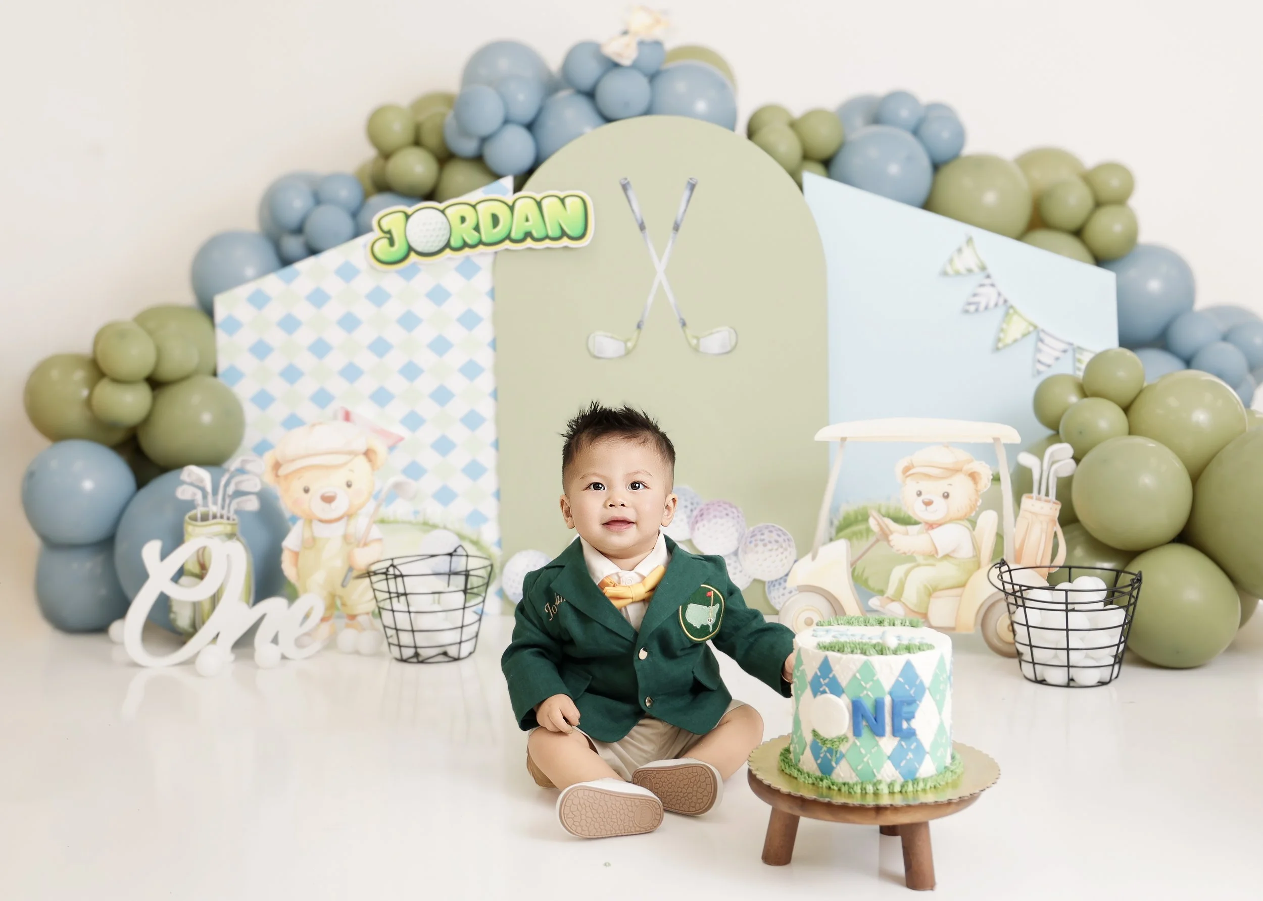 Child in a green blazer sitting on the floor holding a golf-themed birthday cake with the name 'ONE' on it, surrounded by golf-themed decorations and balloons in shades of blue and green.