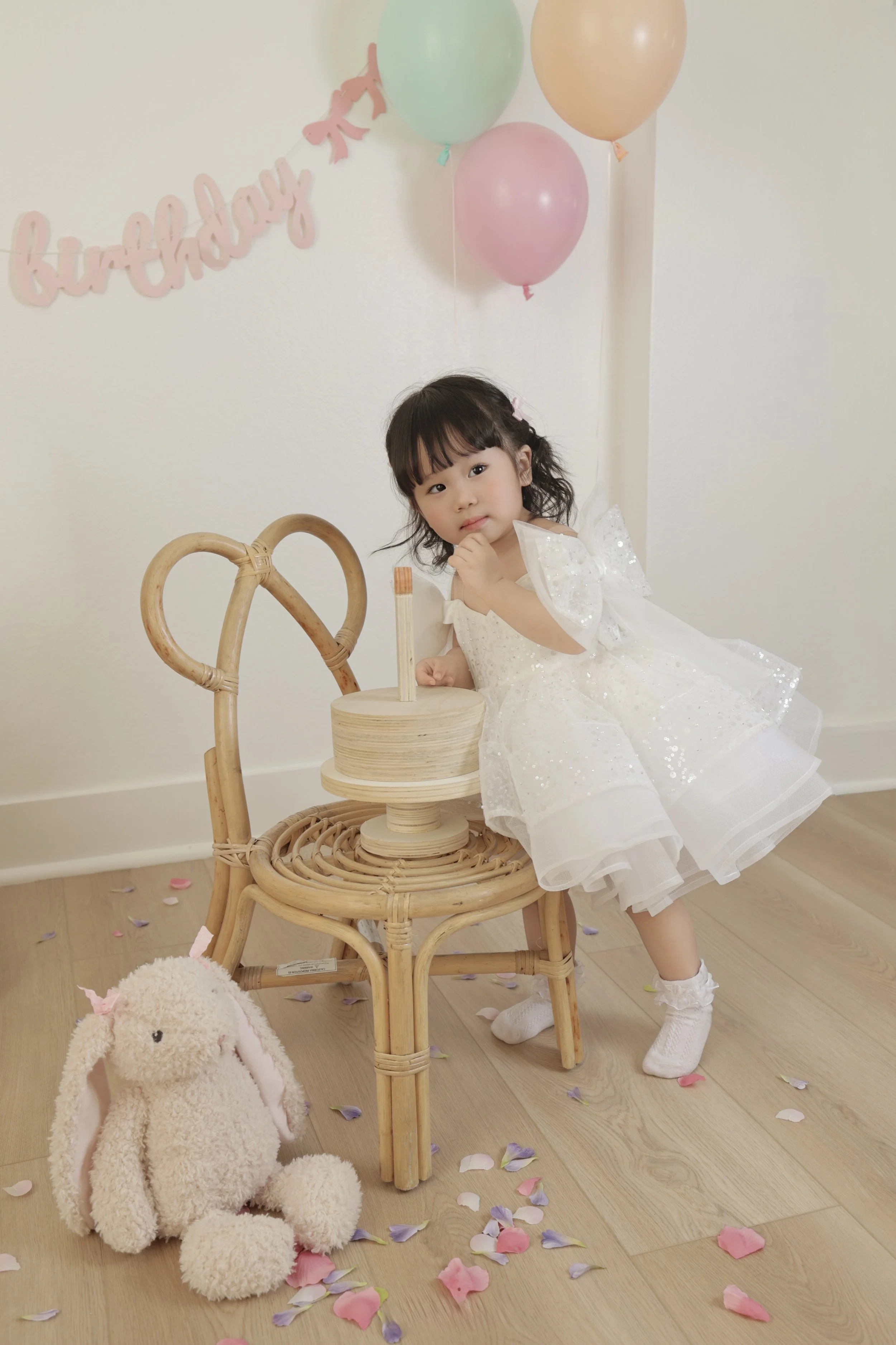 A young girl in a white dress sits next to a wicker chair, posing with her hand on her chin. In front of her on the floor is a plush bunny, and behind her are pink, green, and beige balloons with a pink 'birthday' banner hanging on the wall.