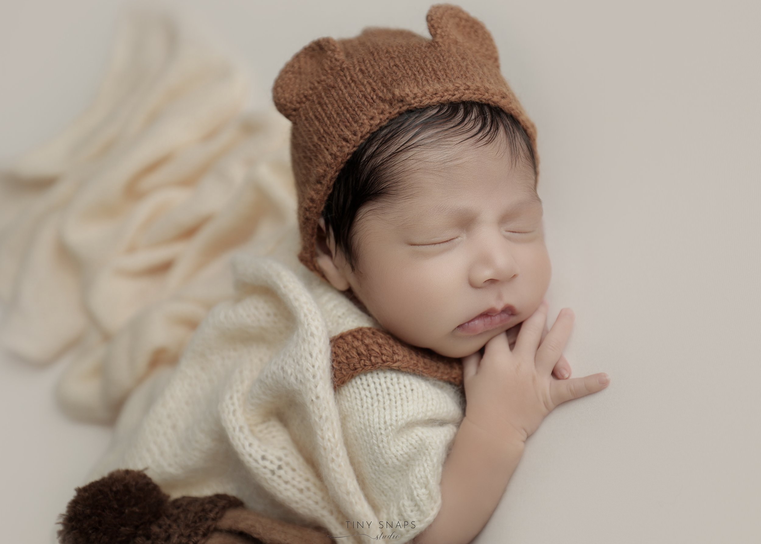 A sleeping newborn baby dressed in knitted cream and brown clothing, including a brown hat with bear ears, resting on a soft cream blanket.