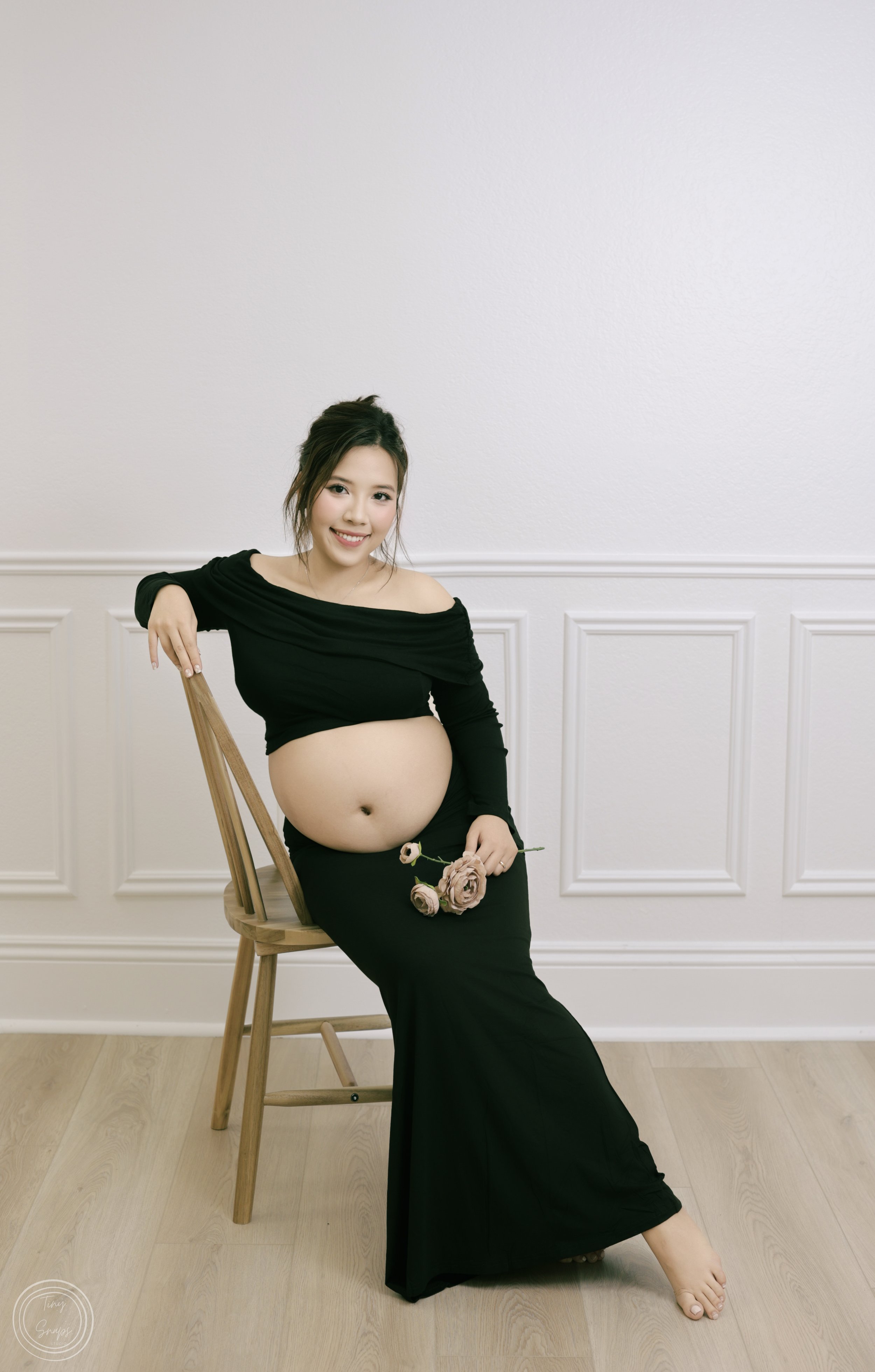 A pregnant woman in a black off-shoulder dress sitting on a wooden chair, smiling, holding three roses, with a light-colored wall and wooden floor in the background.