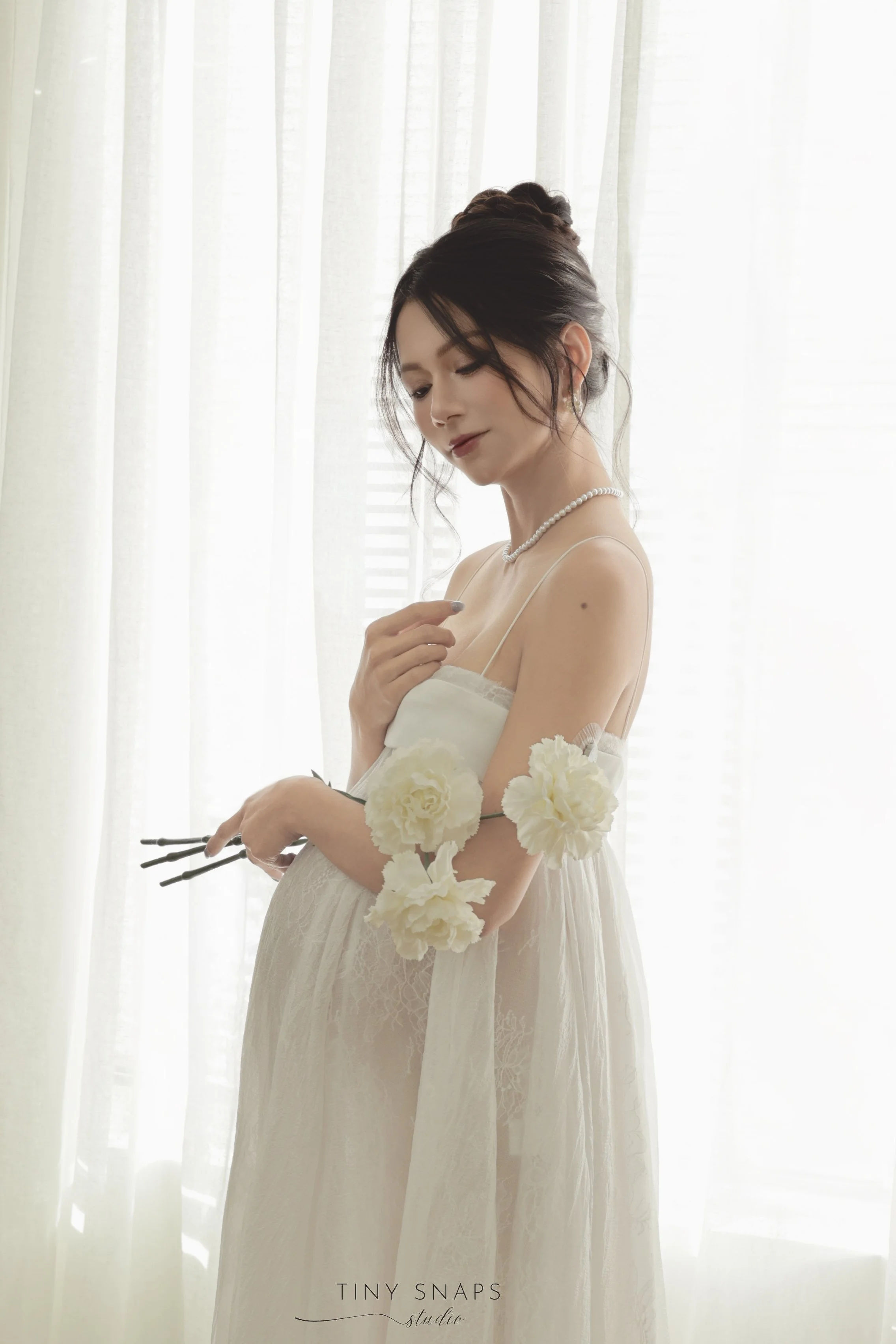 A woman dressed in a white gown with floral accents on her arms, standing near a window with sheer curtains, holding black sticks, and wearing a pearl necklace.