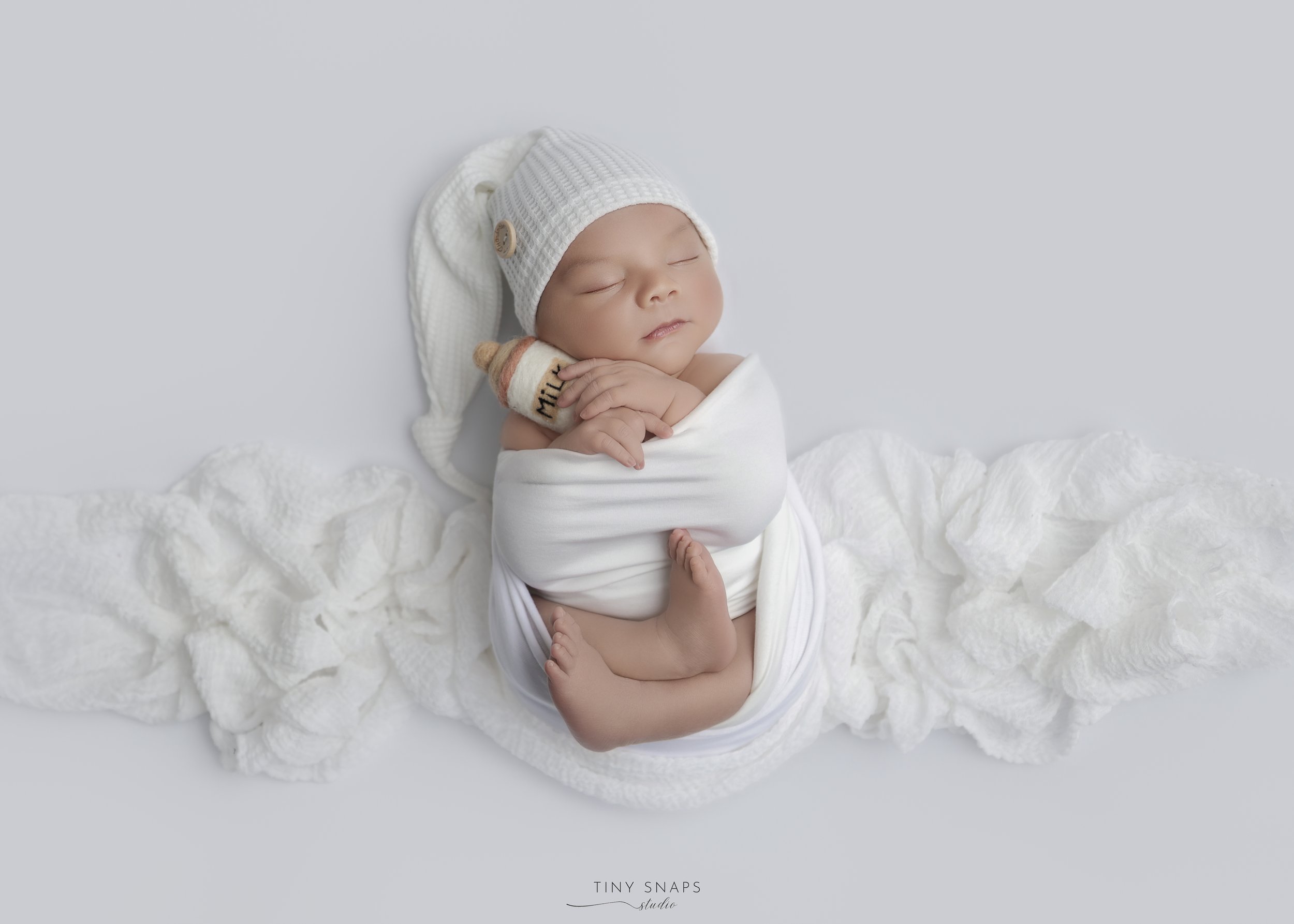 A newborn baby sleeping, wrapped in white fabric, wearing a white knit hat, with a small plush milk bottle, lying on crumpled white fabric on a plain white background.