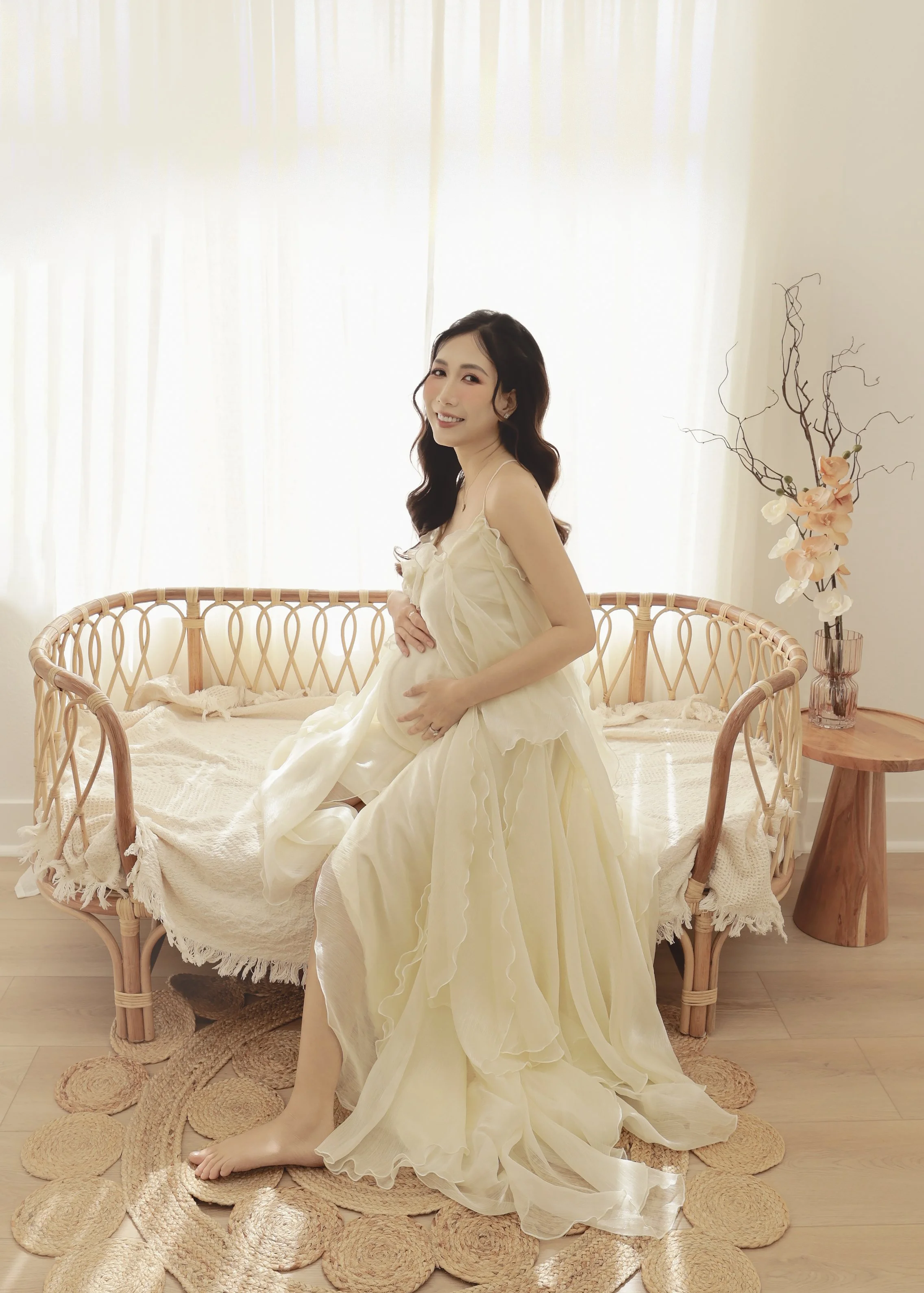 A pregnant woman in a flowing cream-colored dress sitting on a rattan sofa in a bright room with sunlight coming through white curtains, holding her belly and smiling.