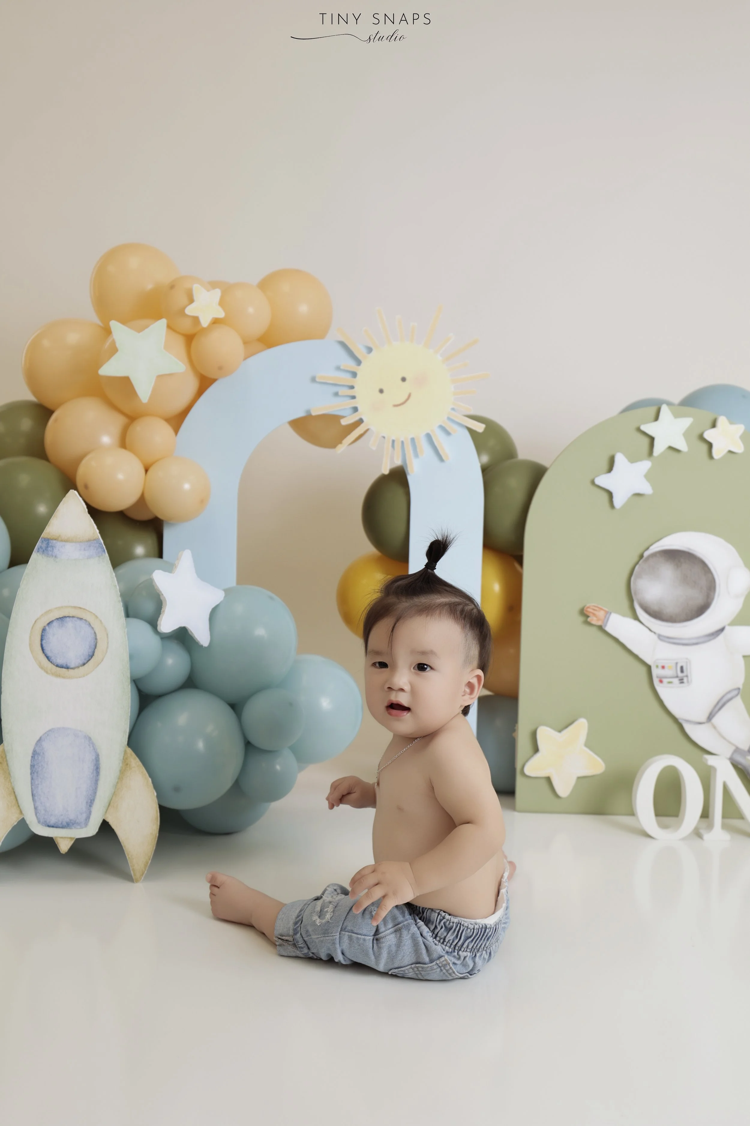 A baby sitting on the floor in front of a space-themed backdrop with balloons and decorations, including a rocket, planets, stars, a sun, and an astronaut.