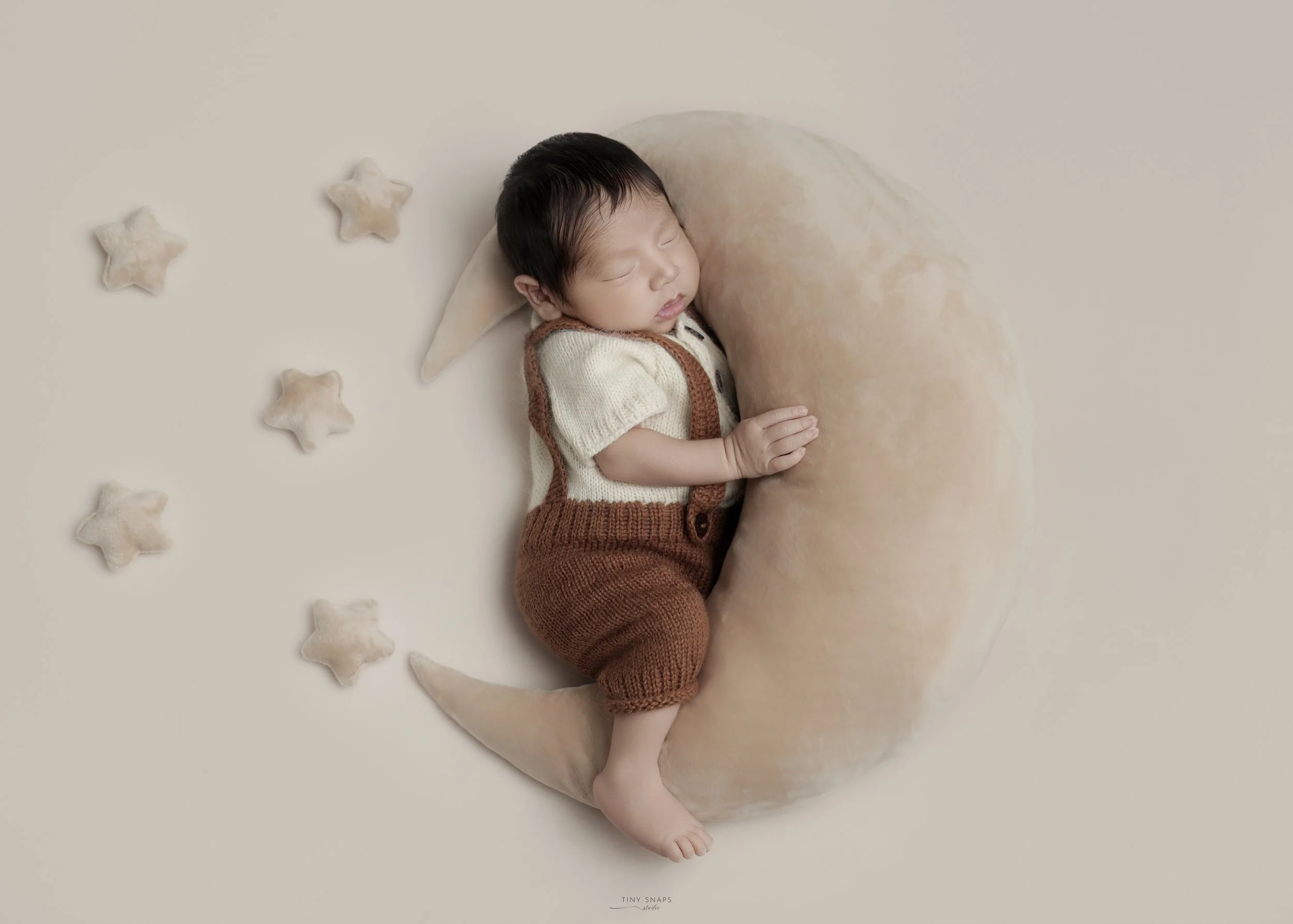 A sleeping baby lying on a crescent moon pillow, surrounded by star-shaped pillows, on a neutral background.