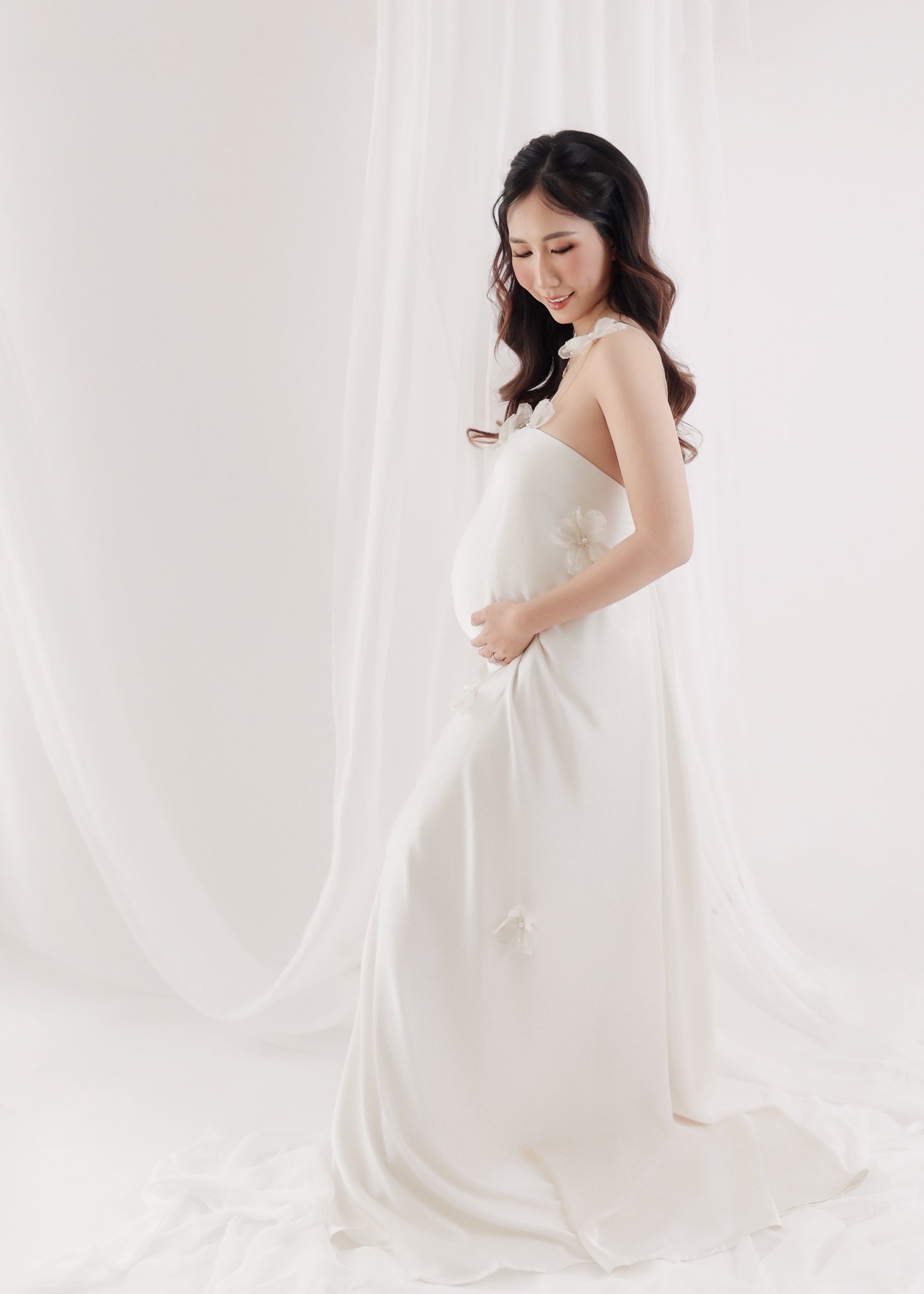 A pregnant woman in a white wedding dress standing with her hands on her belly, looking down and smiling softly, with white curtains in the background.