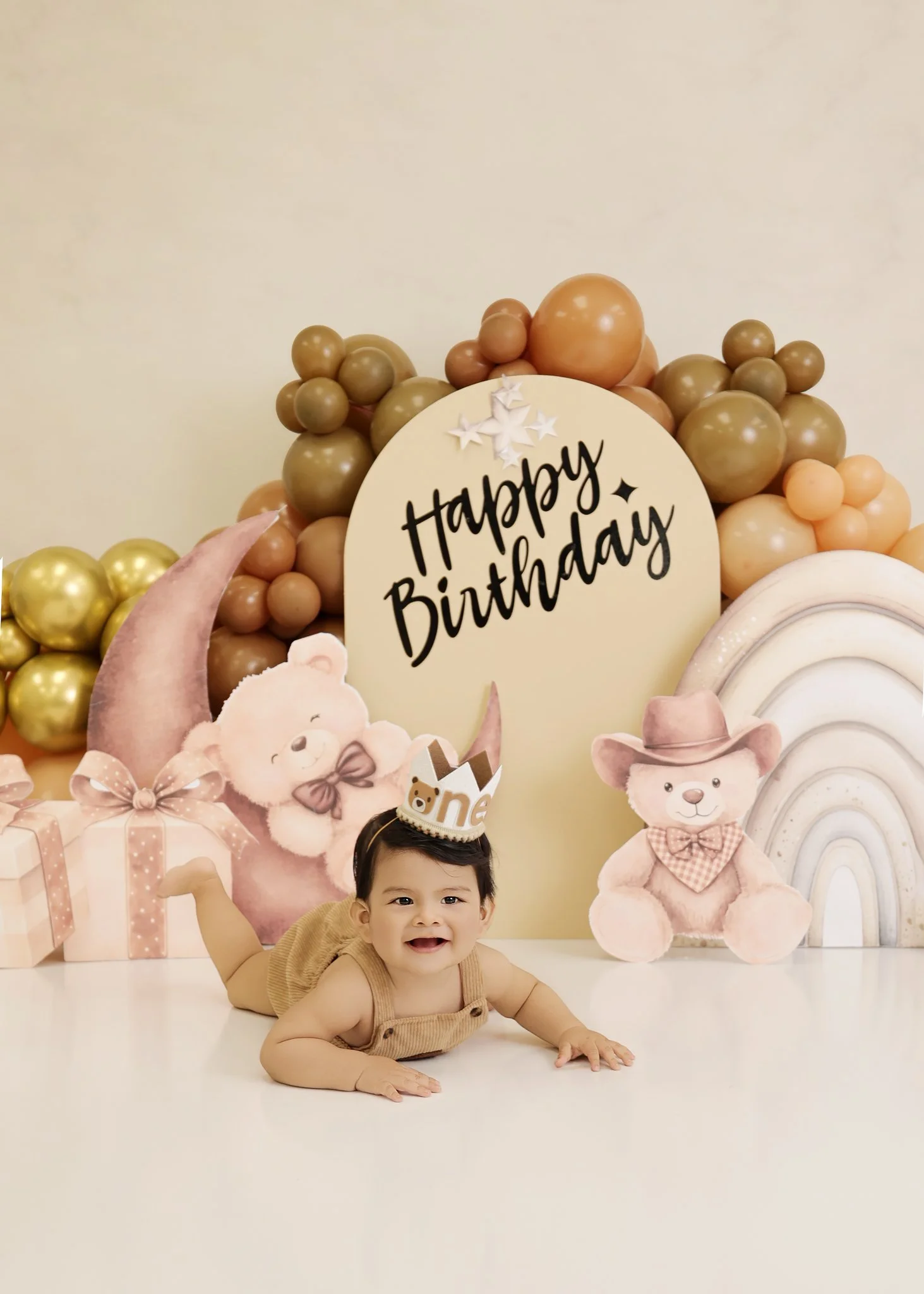 Baby lying on the floor celebrating a birthday with a "Happy Birthday" sign, a birthday crown, bears, presents, and pastel-colored balloon decorations.
