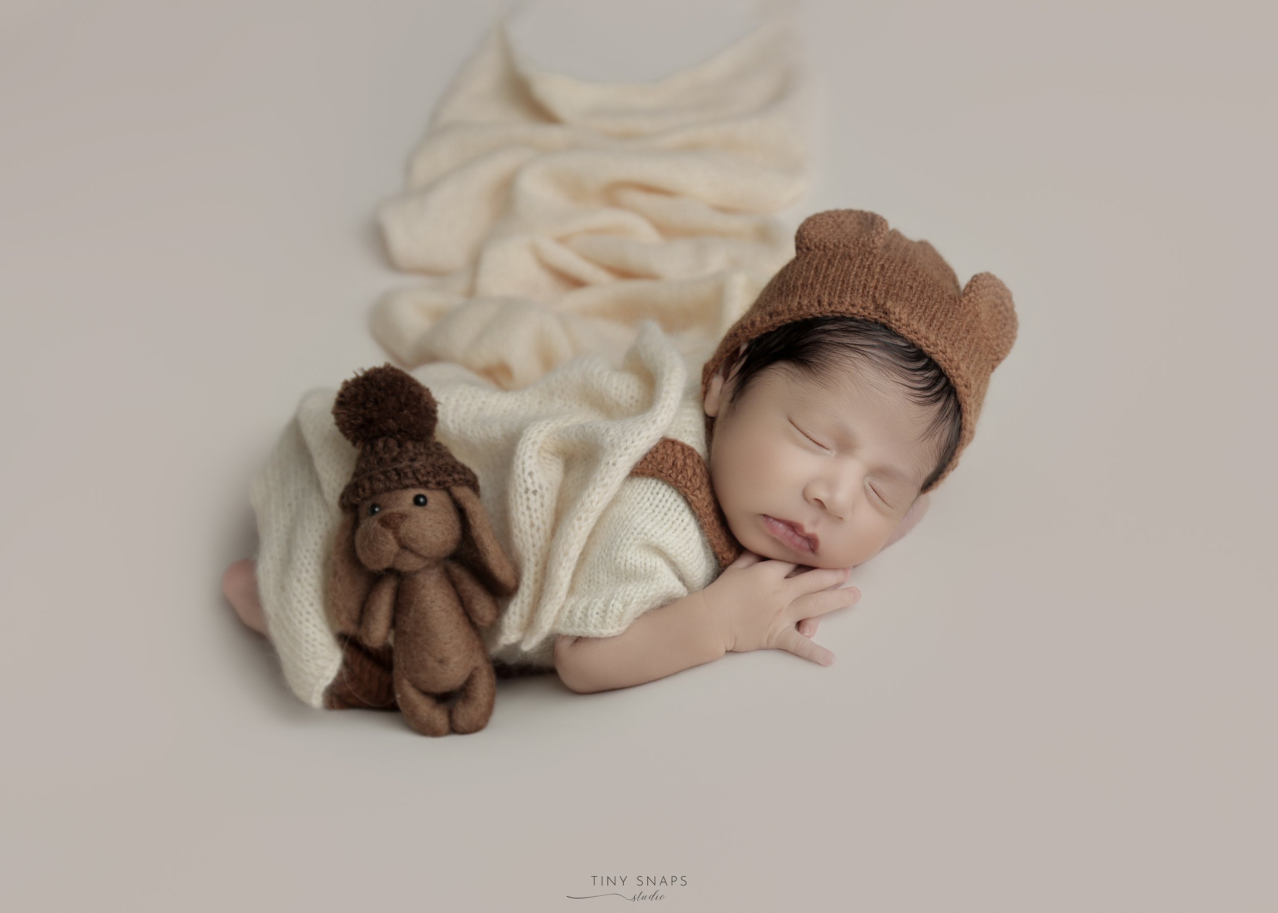 A sleeping baby wearing a brown bear hat and cream-colored outfit, lying on a surface with a cream blanket, with a brown plush bunny toy nearby.