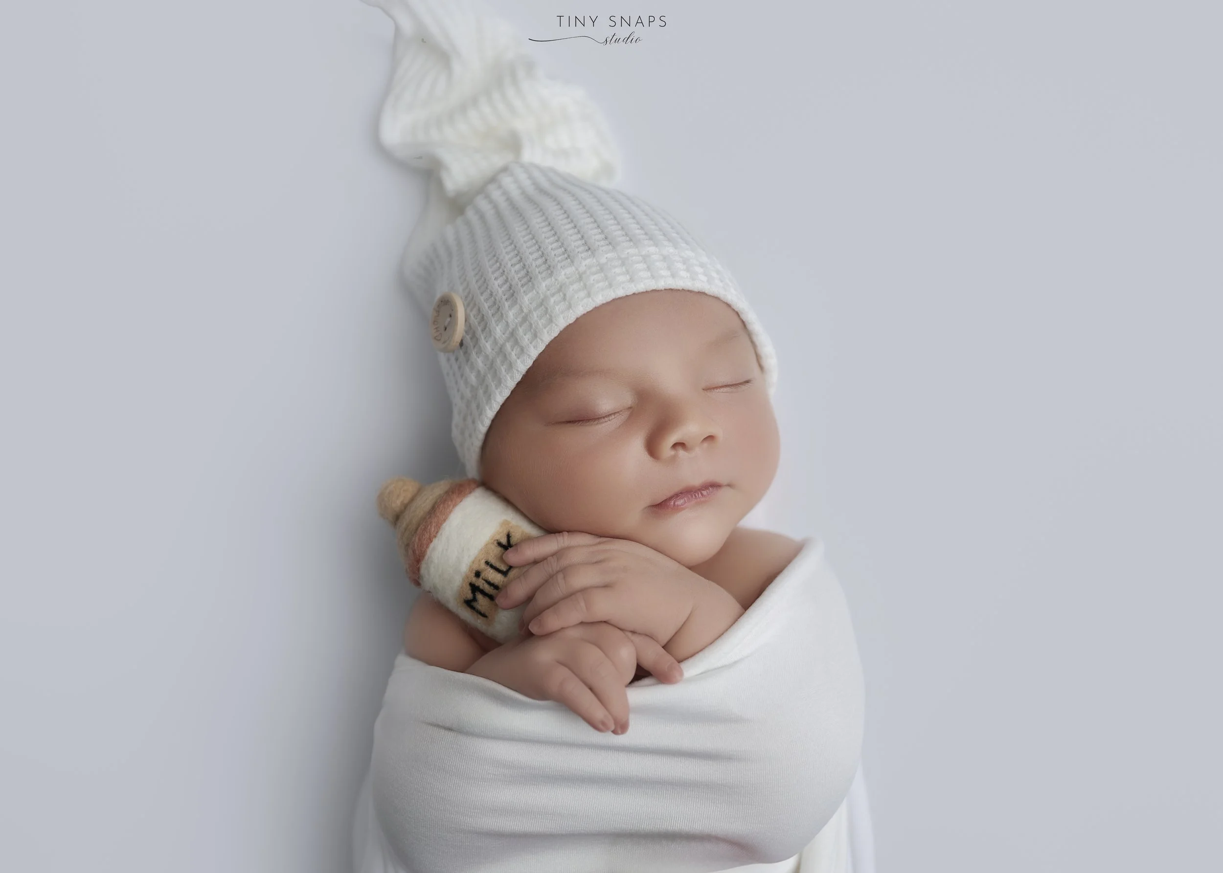 A sleeping newborn baby wrapped in a white blanket, wearing a white knit hat with a long tail, and holding a small plush toy with "Milky" embroidered on it.